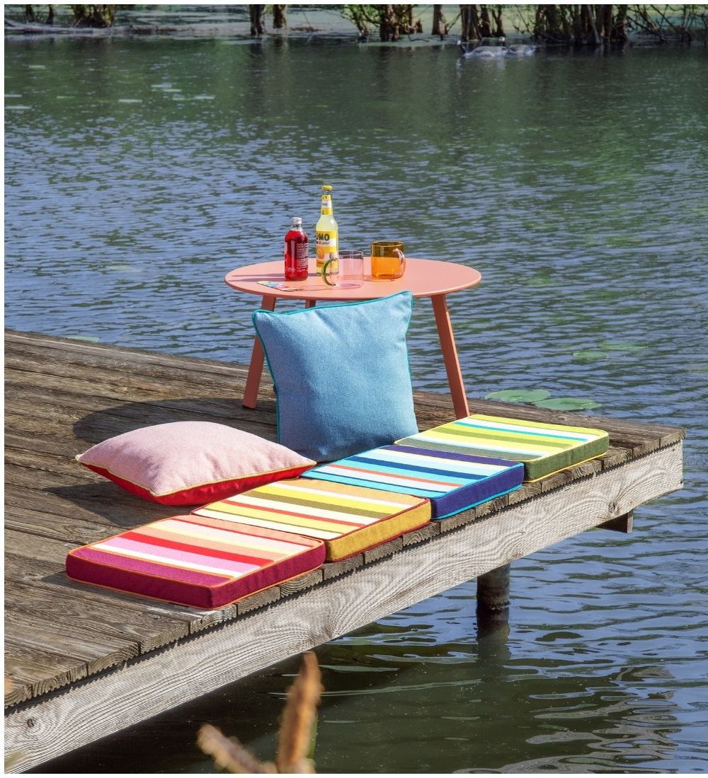 Gemütlicher Sitzbereich auf Holzsteg über ruhigem Wasser. Auf kleinem, rundem lachsfarbenem Tisch stehen Getränke und Gläser. Bunte Kissen auf dem Steg: hellblau, drei gestreift, eins rosa. Pflanzen im Hintergrund.