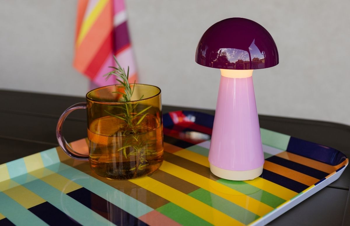 Lampe champignon violette au pied rose pâle sur plateau rayé vif. À côté, verre ambre avec anse violette, rempli d'eau et d'une branche de romarin. Rayures jaunes, vertes, bleues, orange. Tissu rayé au mur.