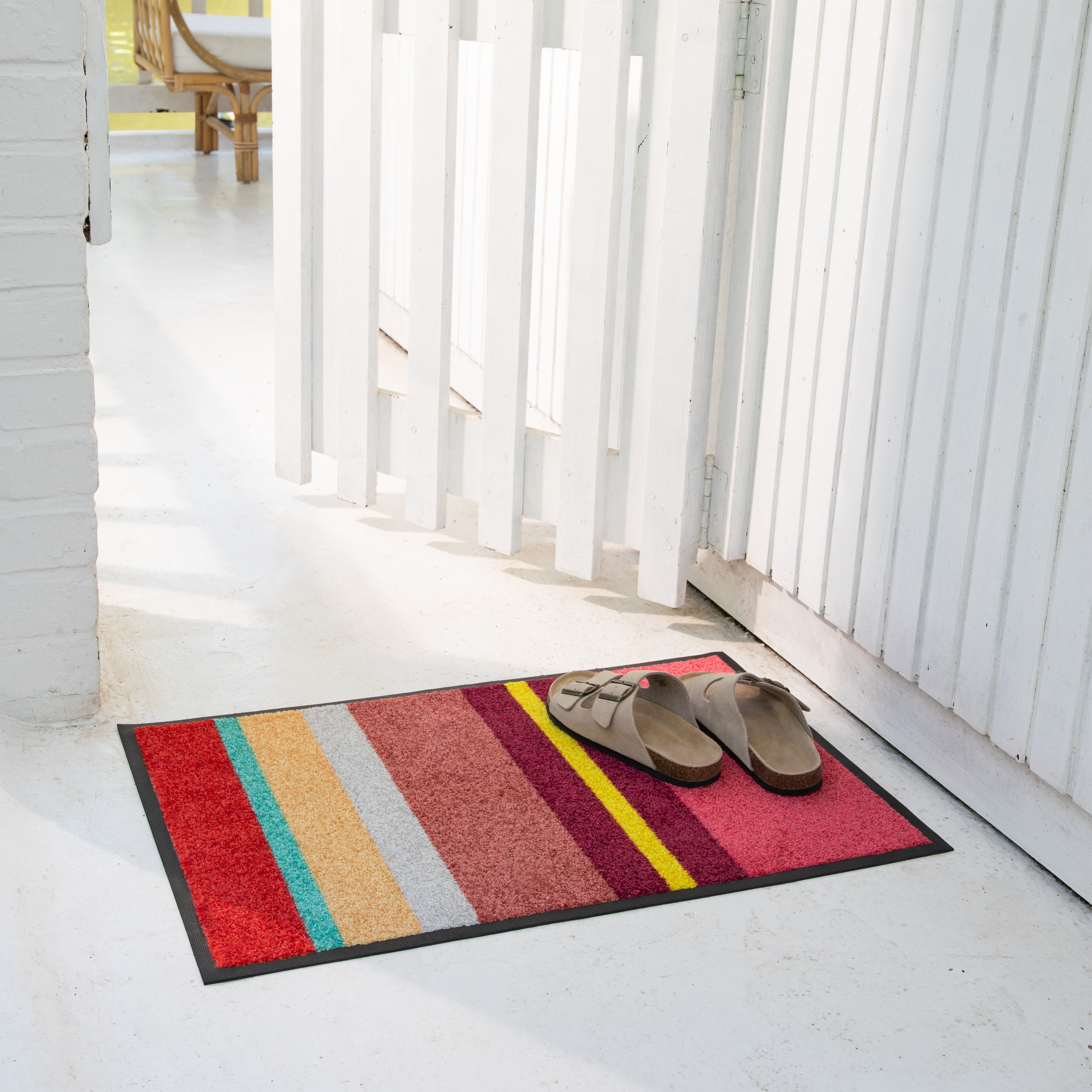 Ein farbenfroher gestreifter Fußabtreter mit Streifen in Rot, Türkis, Orange, Grau, Pink, Gelb und Burgunderrot liegt auf hellem Boden. Darauf steht ein Paar beige Riemensandalen. Rechts ein weißer Holzzaun, links eine weiße Ziegelwand.