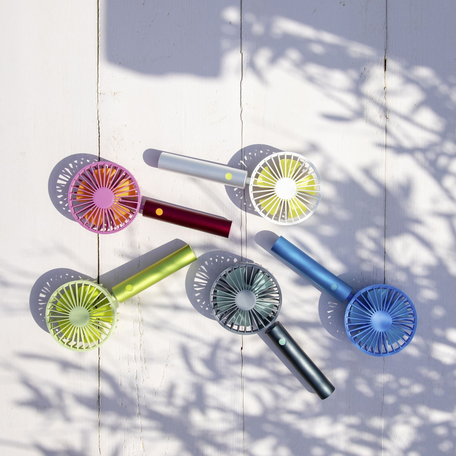 Fünf farbige, aufladbare Akku-Ventilatoren auf hellem Holztisch. Die Farben sind Pink, Silber, Dunkelrot, Grün und Blau. Schatten von Blättern im Hintergrund.