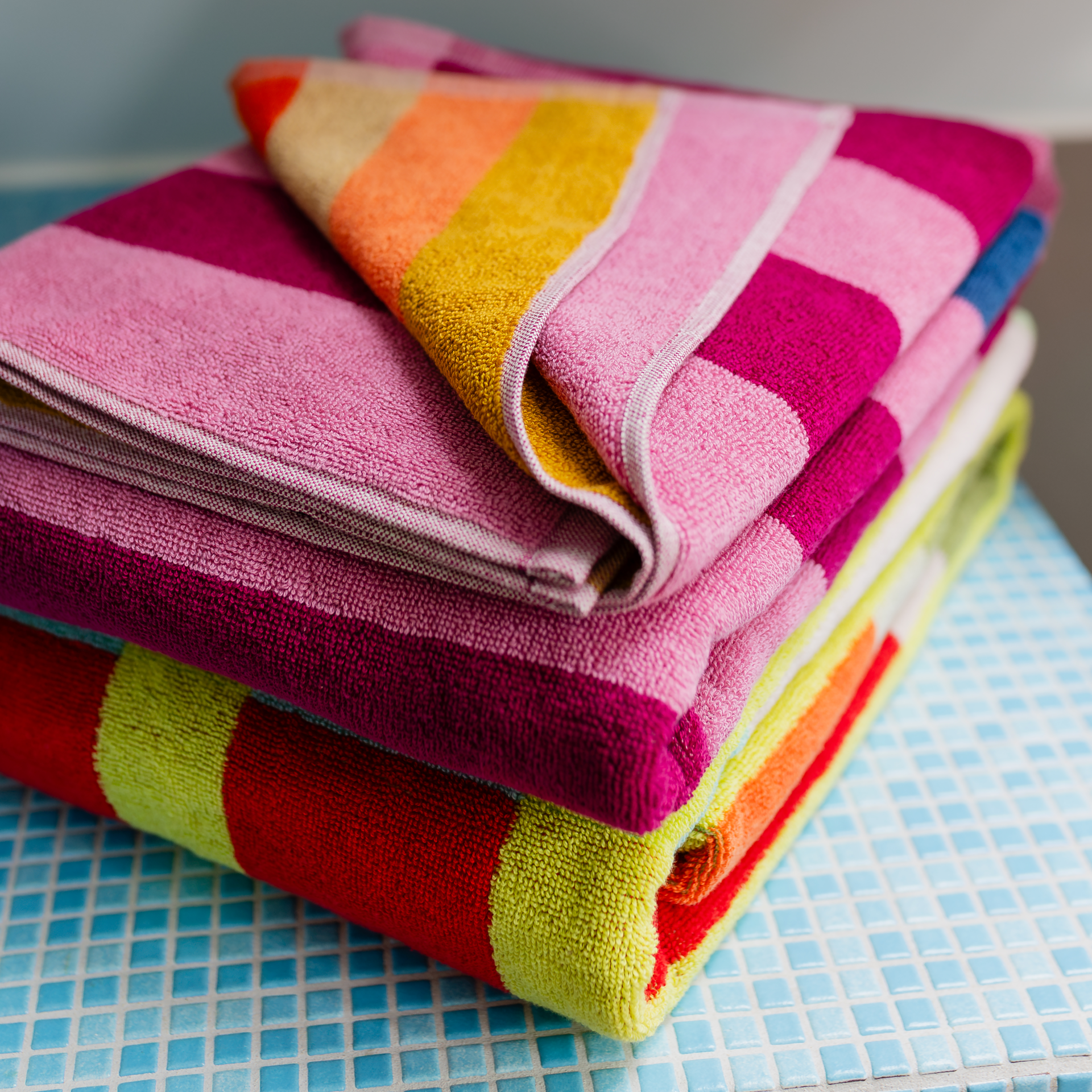 Trois serviettes de bain colorées et rayées sont posées sur une table en mosaïque bleu clair. Moelleuses, roses, jaunes, orange, rouge et vert citron, elles sont bien pliées ; celle du haut est partiellement dépliée et absorbante.
