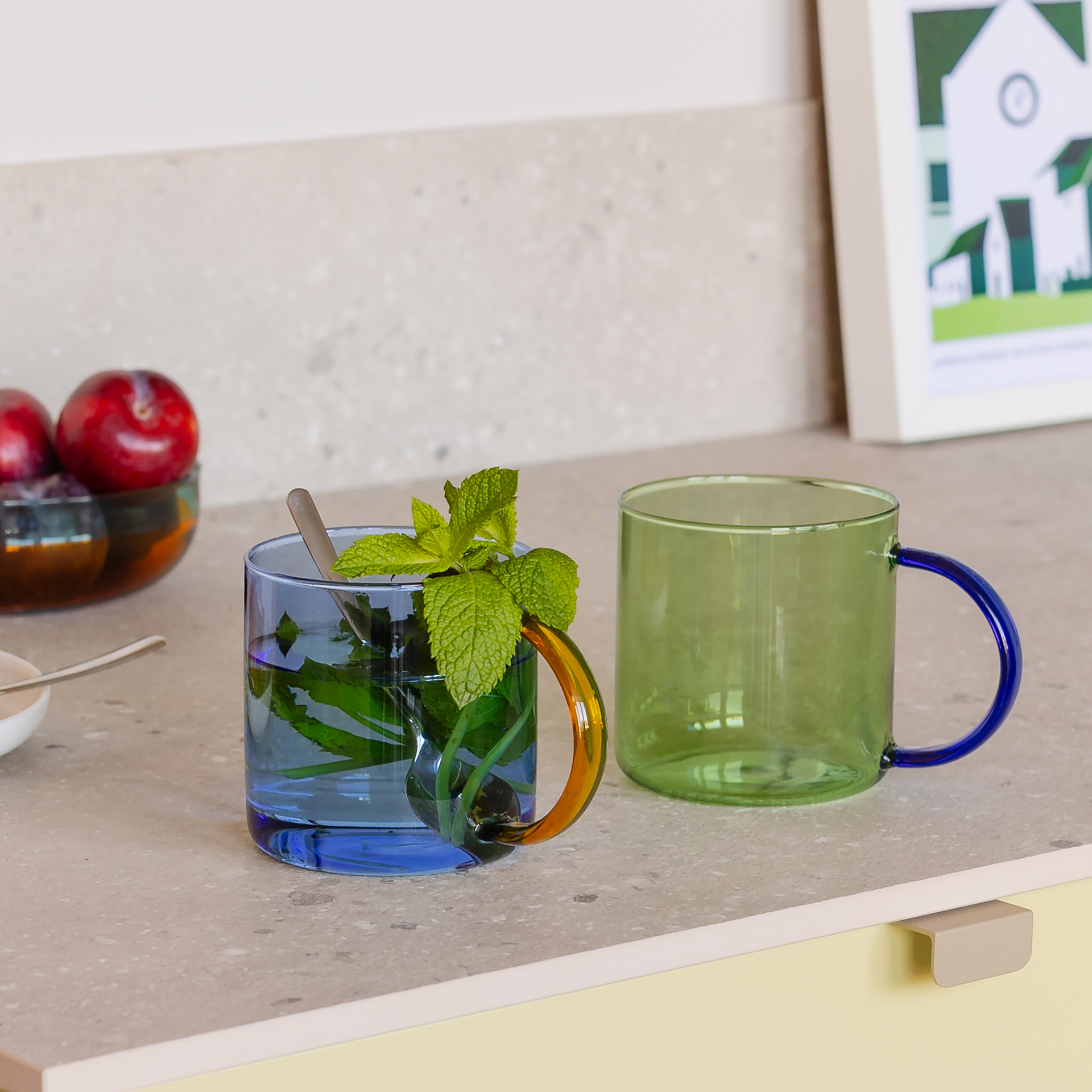 Auf einer hellen Arbeitsfläche stehen zwei farbige Glastassen. In der blauen Tasse steckt ein Löffel und ein Bund Minze. Im Hintergrund stehen Früchte und ein gerahmtes Bild.