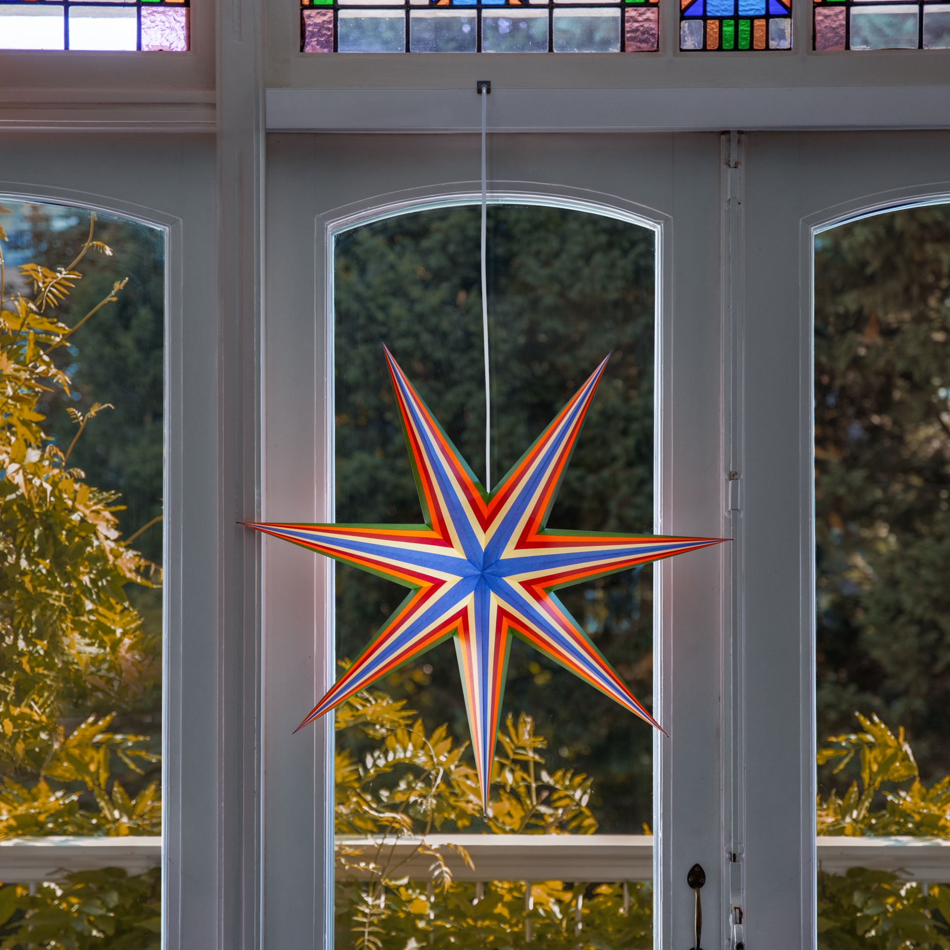 Farbenfroher achtzackiger Papierstern „Mira“ vor weißem Hintergrund. Zacken und Zentrum mit Streifen in Dunkelgrün, Orange, Rot, Beige und Hellblau. Oben weiße Kordel, unten silberner Schnurstopper. Ideal für Fenster/Wände.
