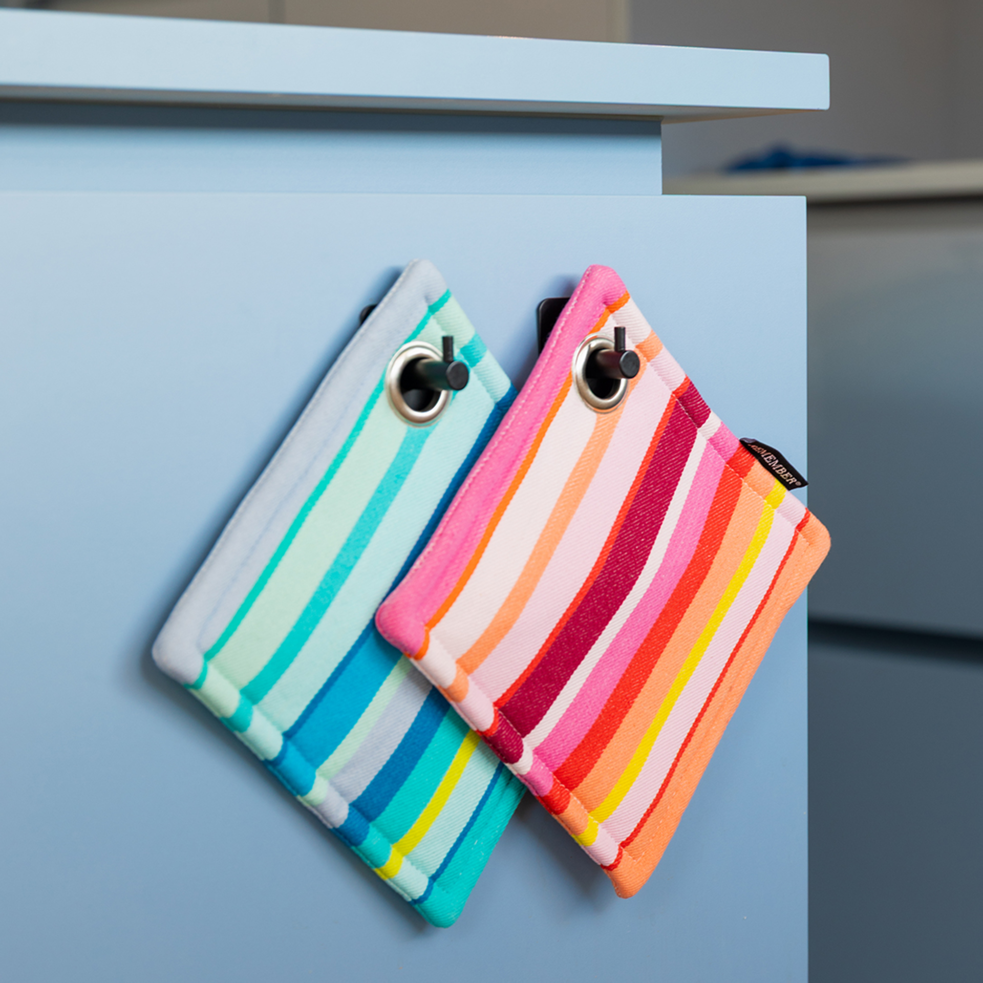 Ein Set bestehend aus zwei gestreiften Topflappen. Einer in Blau- und Grüntönen, der andere in Rosa- und Orangetönen. Beide verfügen über eine Aufhängung mit Metallöse.
