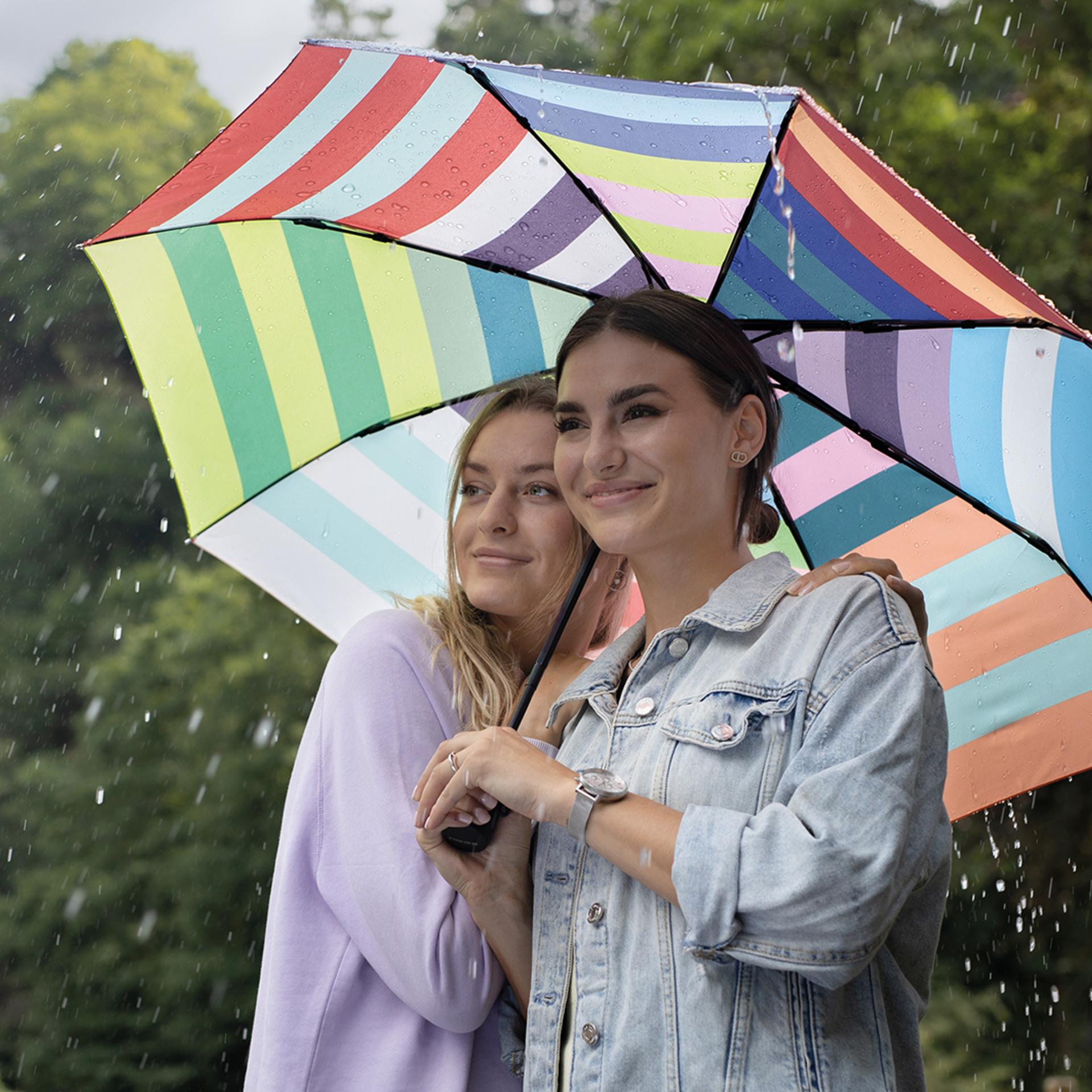 Zwei Frauen unter einem geöffneten, farbenfrohen Taschenschirm im Regen. Der Regenschirm hat ein gestreiftes Muster in verschiedenen Farben.