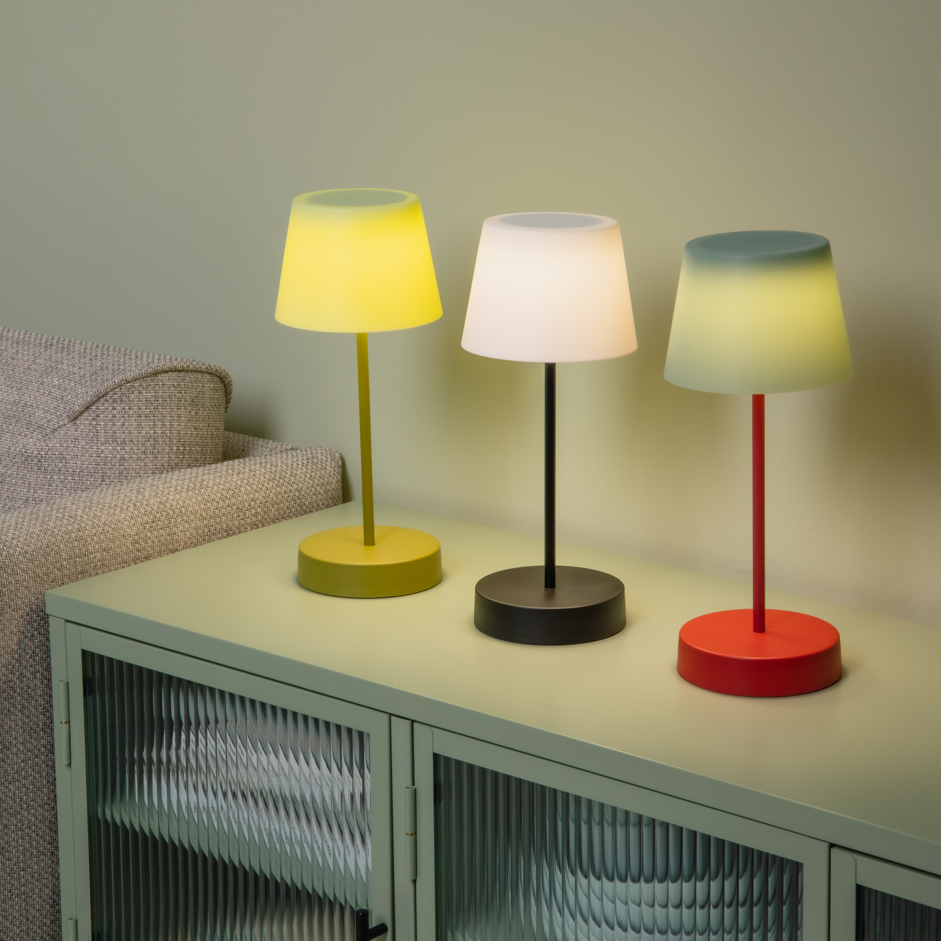 Drei farbige Tischleuchten 'Oscar' stehen auf einem Sideboard: Gelb, Weiß und Rot. Die Lampenschirme sind beleuchtet. Ein Teil eines Sofas ist im Hintergrund erkennbar.