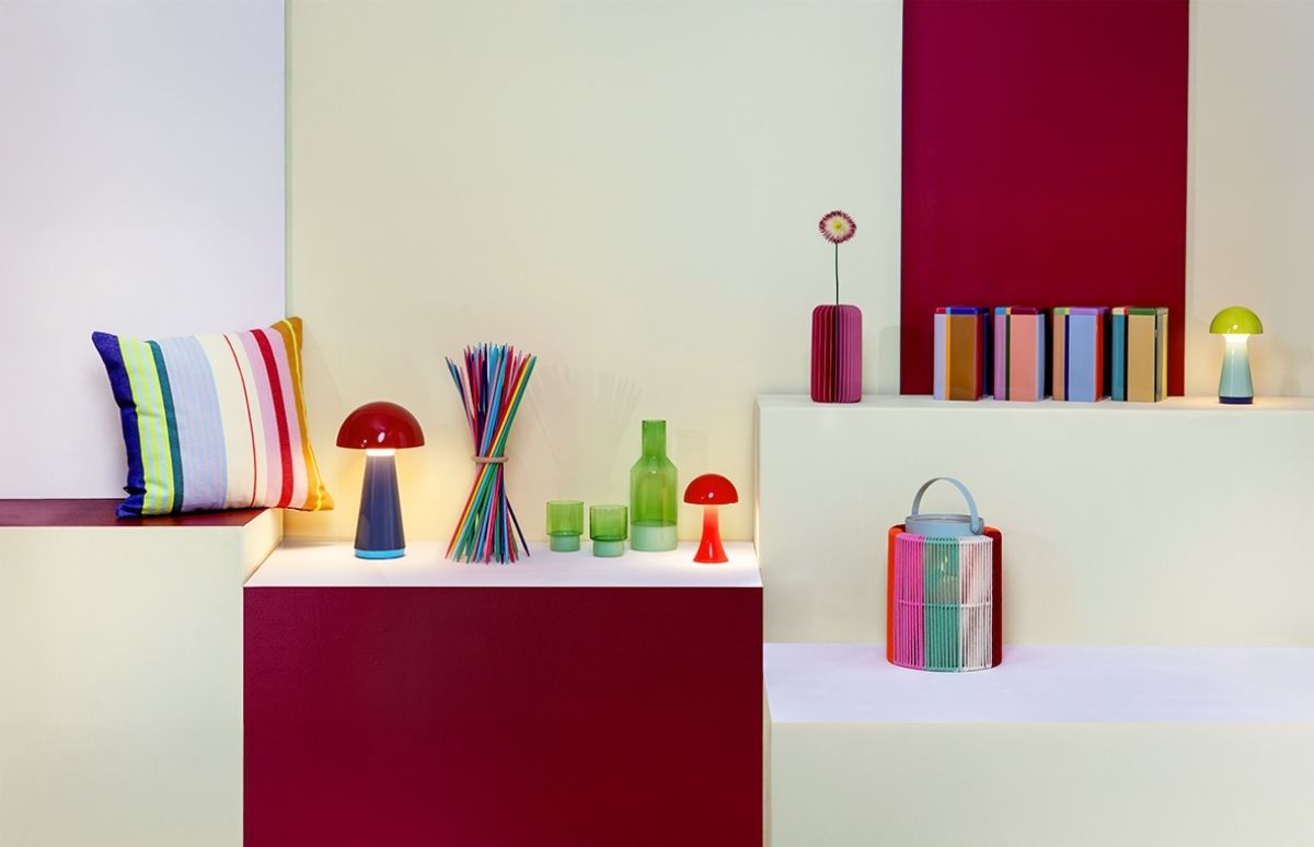 Moderne Präsentation bunter Dekoartikel auf weißen und weinroten Displays: gestreiftes Kissen, rote und grüne Pilzlampen, bunte Trinkhalme, grüne Gläser, pinke Vase mit Blume, gestreifte Blöcke und große Laterne.