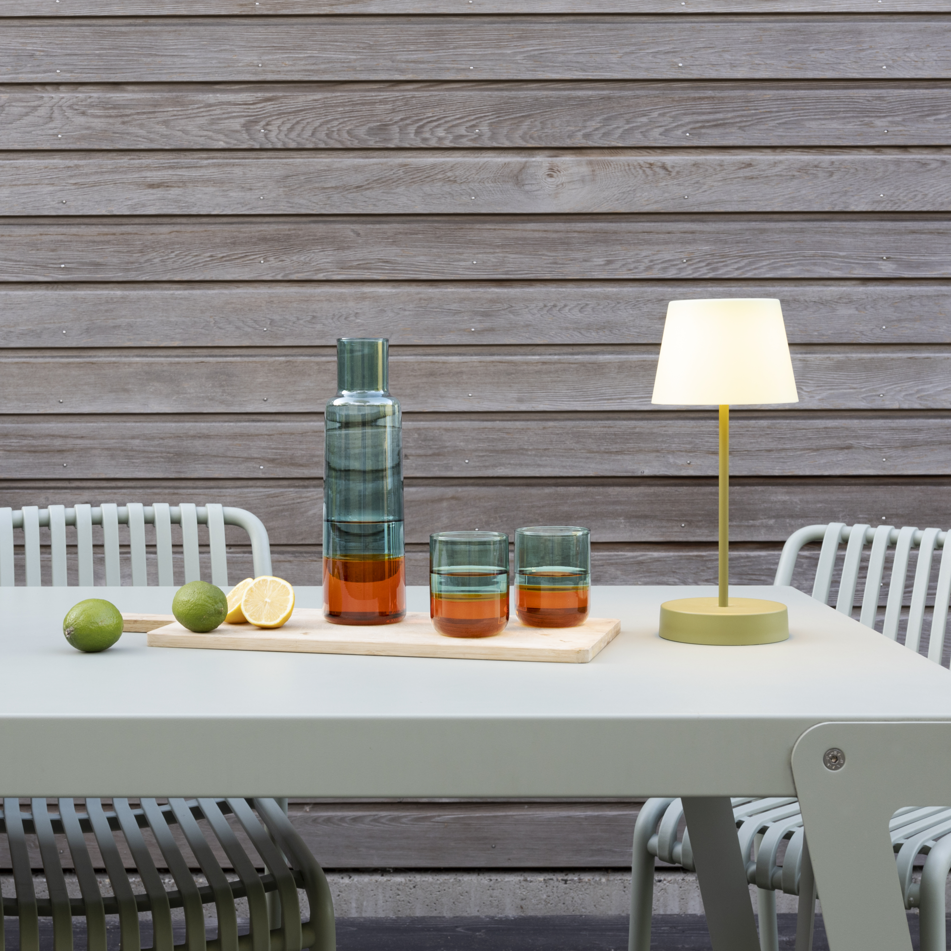 Glaskaraffe 'amber' mit zwei Gläsern und Zitrusfrüchten auf Tisch mit Lampe. Karaffe und Gläser sind zweifarbig, oben grün, unten amberfarben.