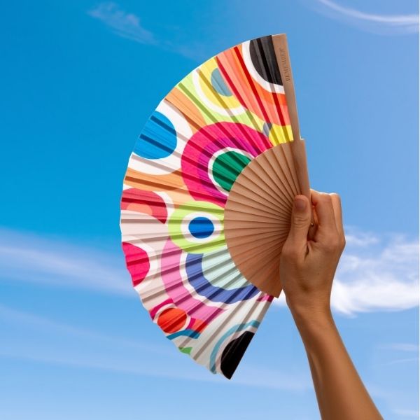 Une main tient un éventail ouvert devant un ciel bleu éclatant parsemé de nuages légers. L'éventail présente un motif vivant composé de cercles et de demi-cercles rouges, jaunes, bleus et roses. Les baleines sont en bois clair.