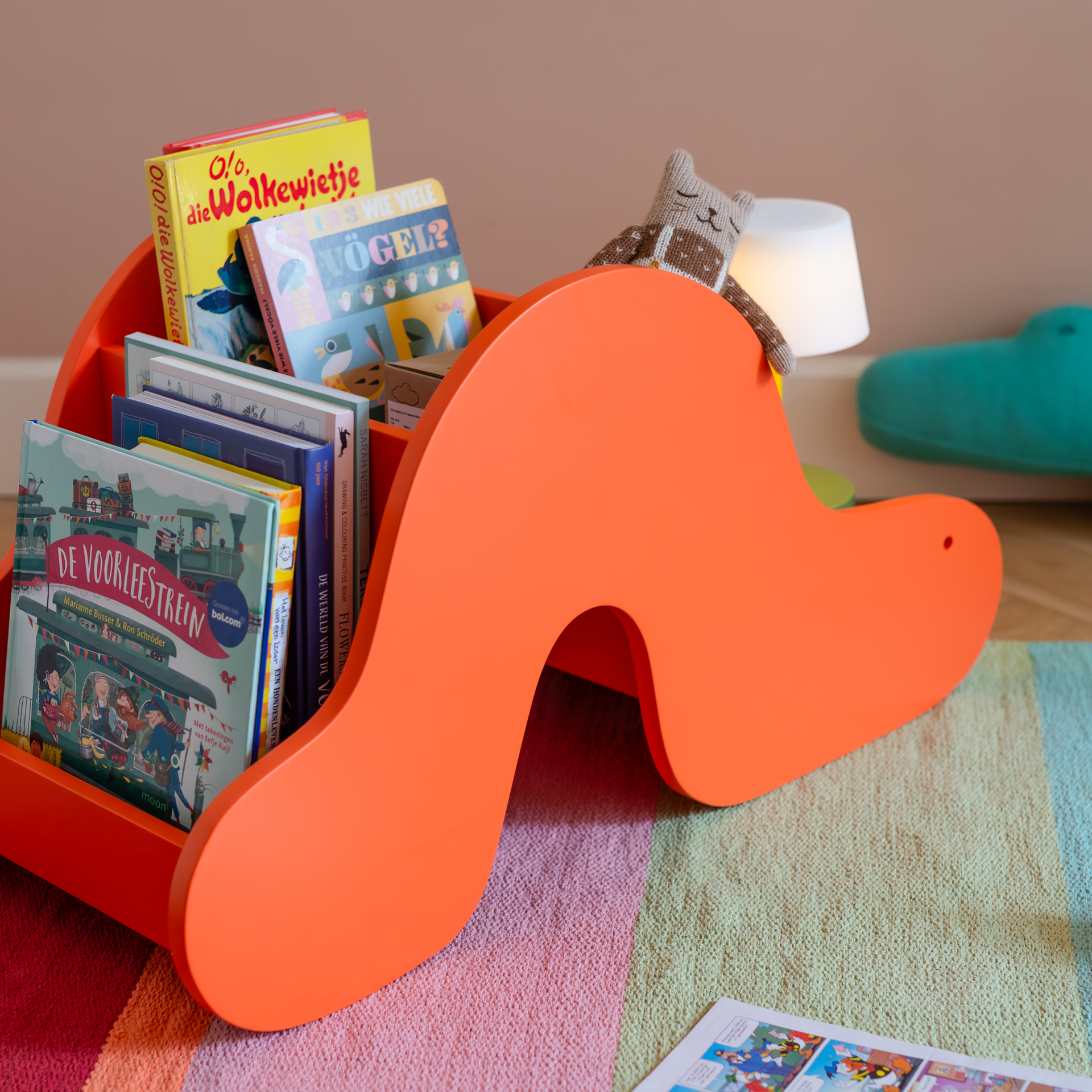 Ein orangefarbenes, wellenförmiges Bücherregal mit Kinderbüchern und einem gehäkelten Stofftier in einem Kinderzimmer auf einem gestreiften Teppich.