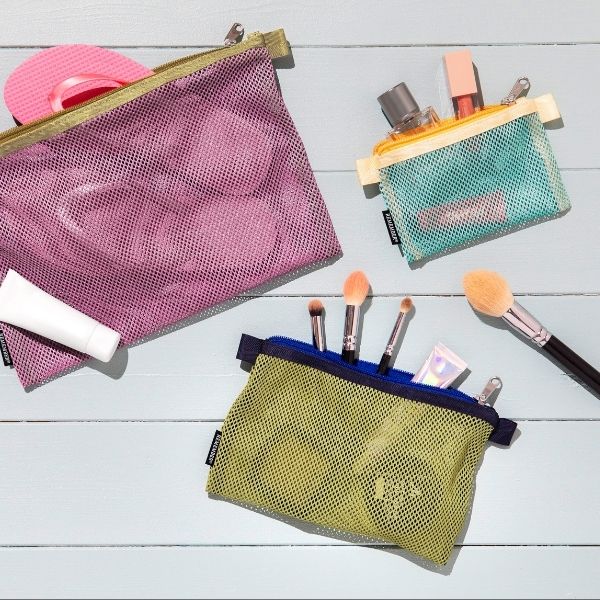 Trois pochettes en filet colorées avec articles de voyage et beauté sur fond bois bleu clair. Violette : tongs roses, tube de crème solaire. Turquoise : parfum, gloss. Vert citron : pinceaux maquillage, crème.