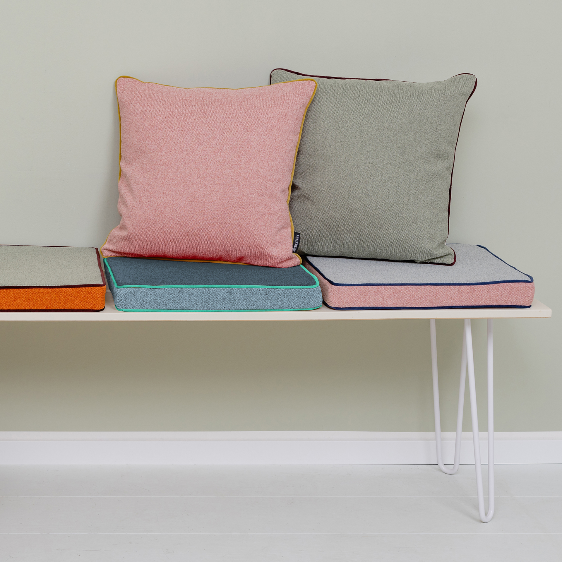 Coussins multicolores avec passepoil sur un banc. Le coussin rose est posé sur le bleu, à côté le gris sur le rose. A gauche, coussin orange. L'arrière-plan est clair.