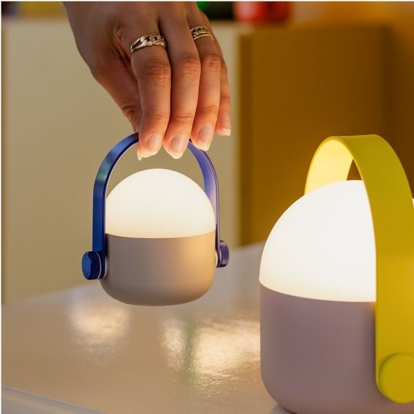Eine Hand hält eine kleine, leuchtende, zylindrische Lampe mit blauem Griff. Sie hat eine graue Basis und einen weißen, diffus leuchtenden oberen Teil. Daneben steht eine ähnliche Lampe mit lila Basis und gelbem Griff.