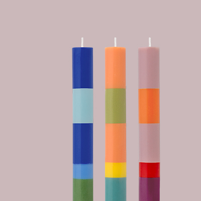 Drei mehrfarbige Stumpenkerzen, von blau über orange bis rosa, im Hochformat auf rosa Hintergrund. Jede Kerze ist in Farbblöcke unterteilt.