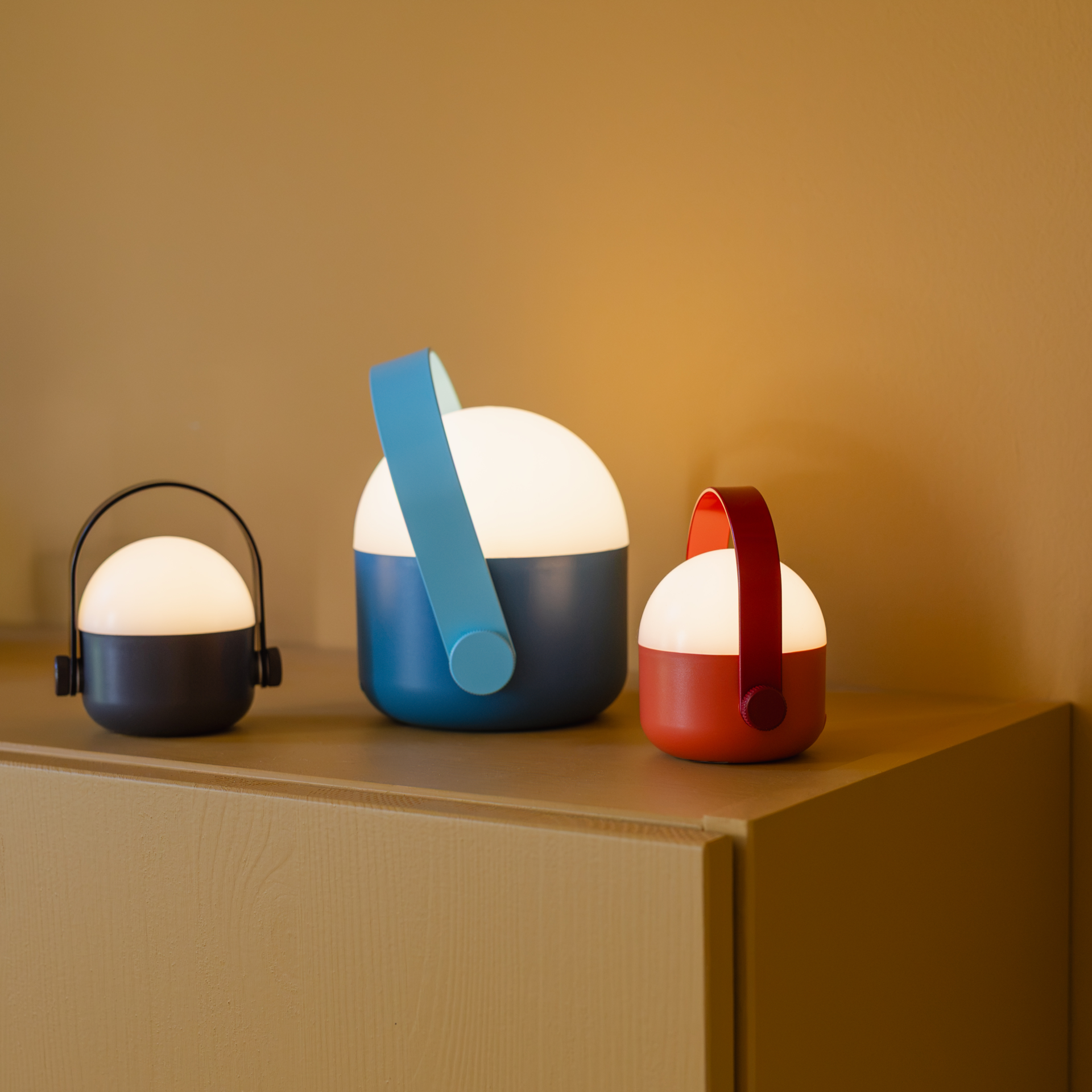 Drei akkubetriebene LED-Leuchten stehen auf einem Holztisch vor gelb-brauner Wand: links grau mit schwarzem Bügel, mittig größer blau mit hellblauem Band, rechts rot mit rotem Band. Oben je weiße Halbkugel, warmweißes Licht.