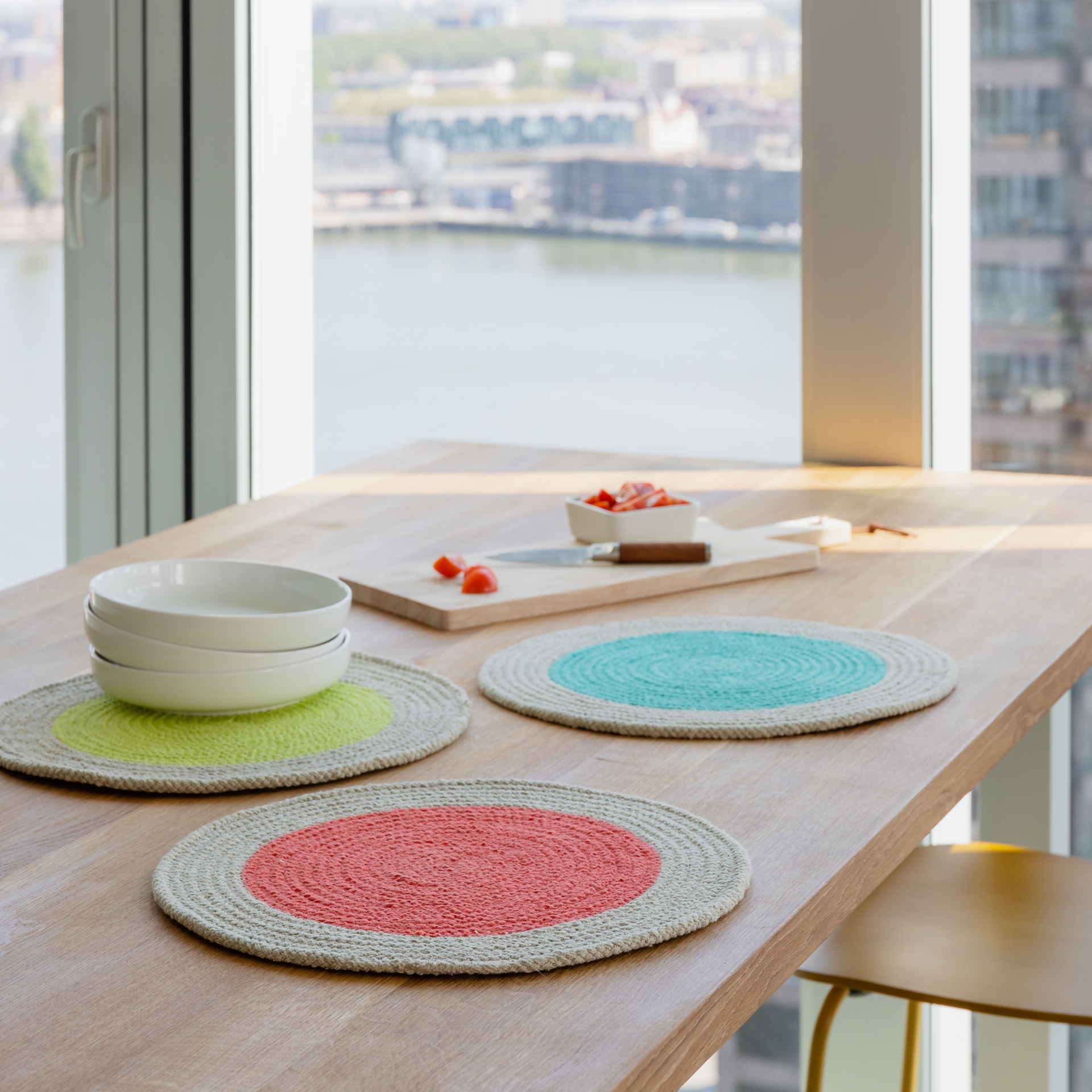 Drei runde Baumwoll-Tischsets in Mint, Rot und Türkis auf einem Holztisch. Geschirr, Schneidebrett mit Tomaten und eine Stuhl sind auch zu sehen. Im Hintergrund eine Fensterfront mit Blick auf die Stadt.