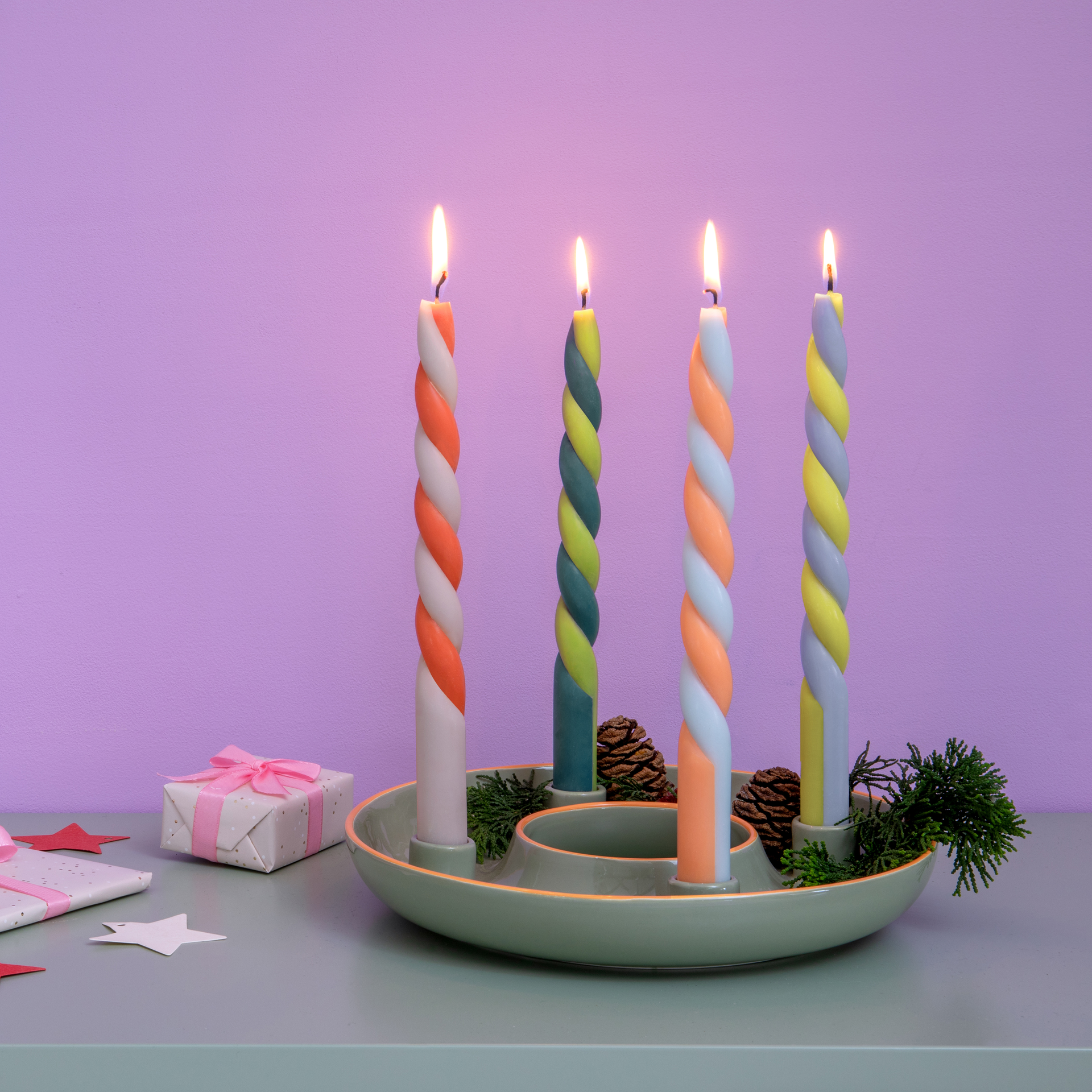 Bougies bâton 'Helsinki' torsadées, colorées et allumées, dans un plateau de bougies vert avec des pommes de pin et des cadeaux sur une table sur fond violet.