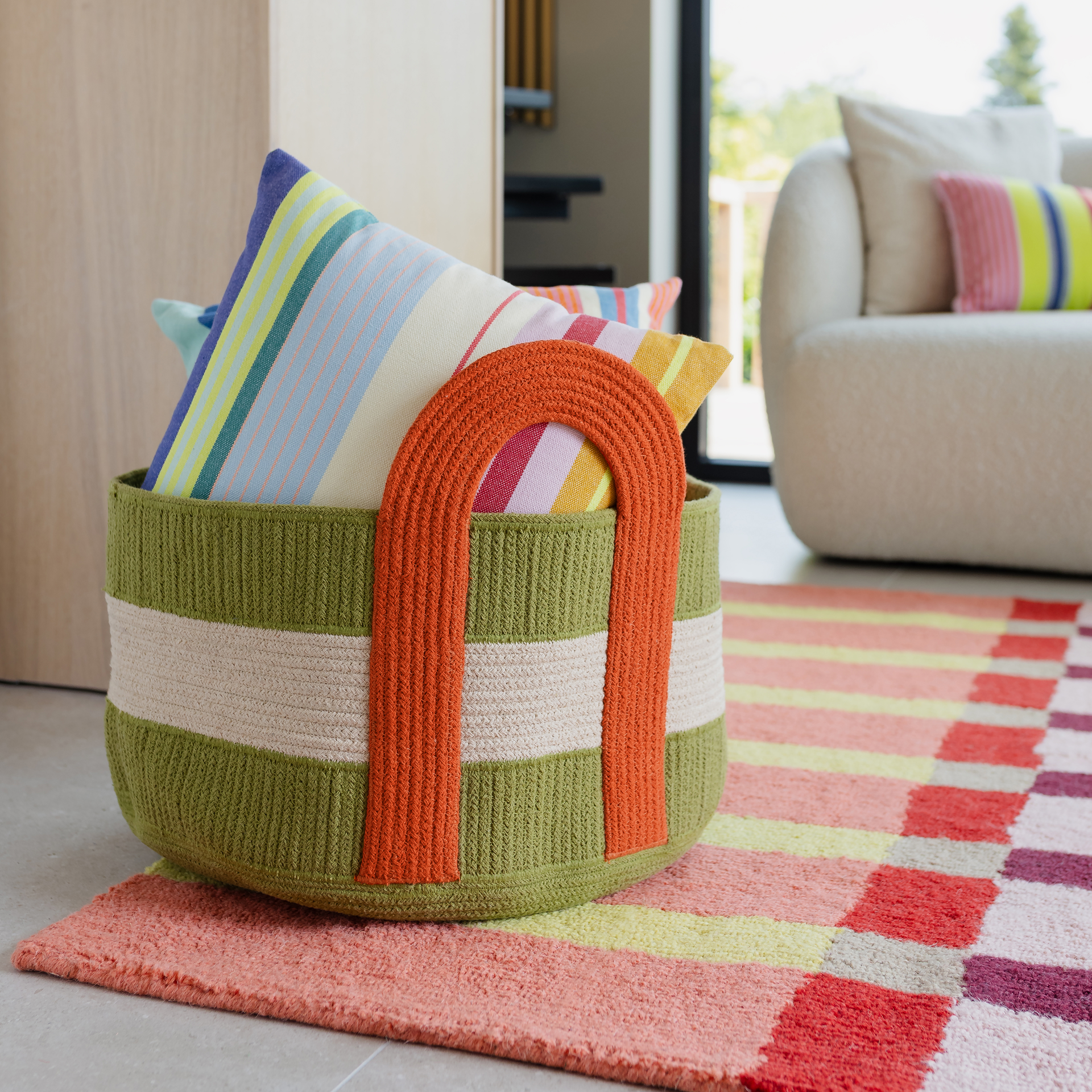 Panier rond et plat vert olive et beige en corde de coton tressée, avec une poignée de transport orange en U caractéristique. À l'intérieur, des coussins rayés multicolores. Posé sur un tapis rayé dans un salon lumineux.