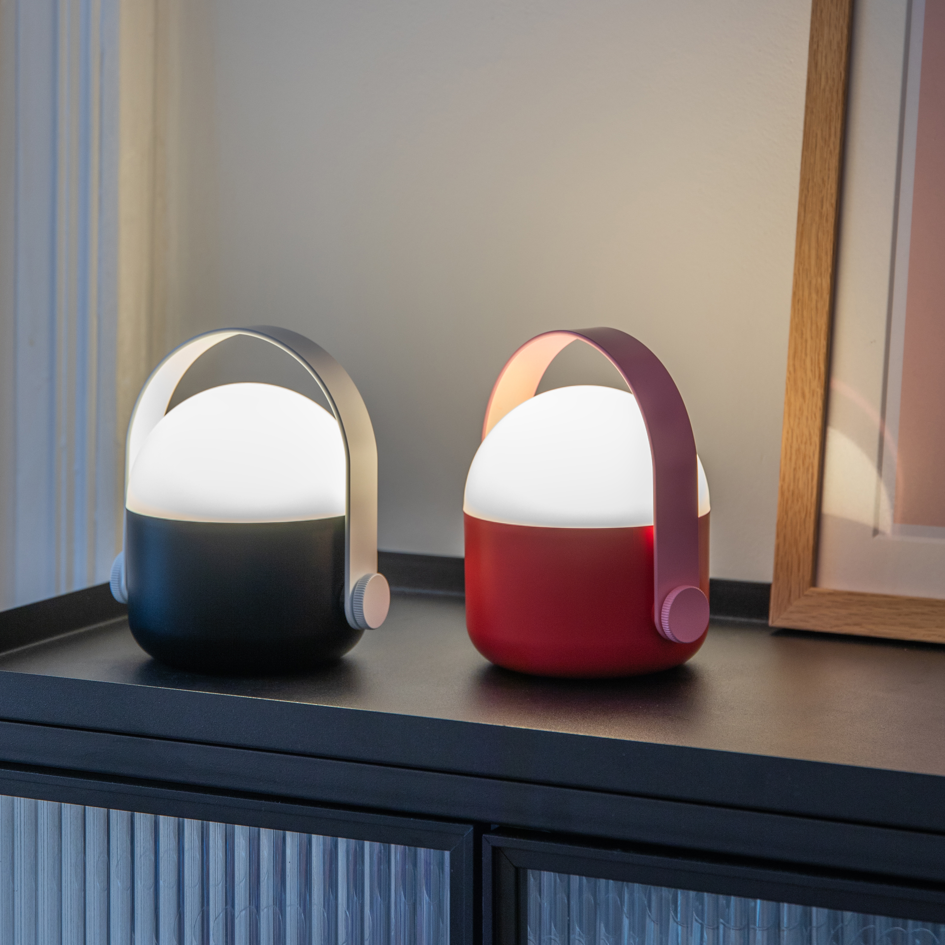 Zwei tragbare Lampen, eine schwarz, eine rot, mit leuchtend weißem Diffusor, stehen auf einem schwarzen Schrank. Im Hintergrund ein Bild mit Holzrahmen.