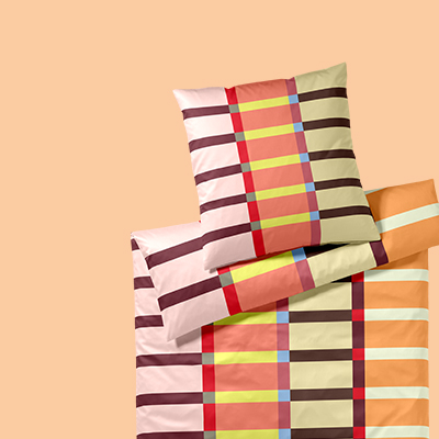 Bettwäsche mit Karomuster in Rosa-, Gelb-, Braun- und Orangetönen auf einem hellen, einfarbigen Hintergrund. Kissen liegen versetzt auf einer Bettdecke.