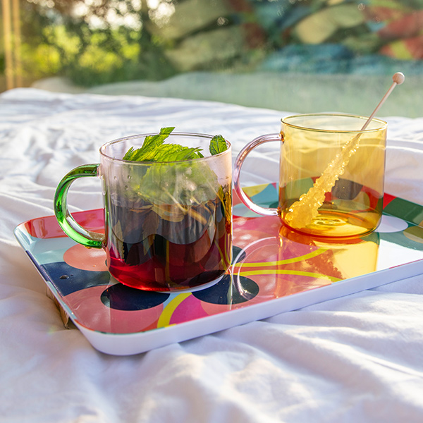 Klarer Glasbecher mit frischen Minzblättern in dunkelrotem Tee neben einem bernsteinfarbenen Glasbecher mit goldenem Kandisstab. Beide auf bunt gemustertem Tablett auf zerknitterten Bettlaken, Sonnenlicht vom Fenster.