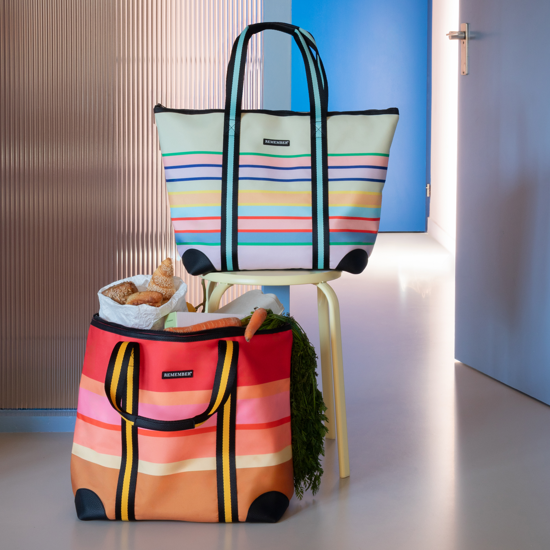 Zwei bunte Einkaufstaschen, gestreift, mit Lebensmitteln gefüllt. Die obere Tasche steht auf einem Hocker, eine Tür ist im Hintergrund sichtbar.