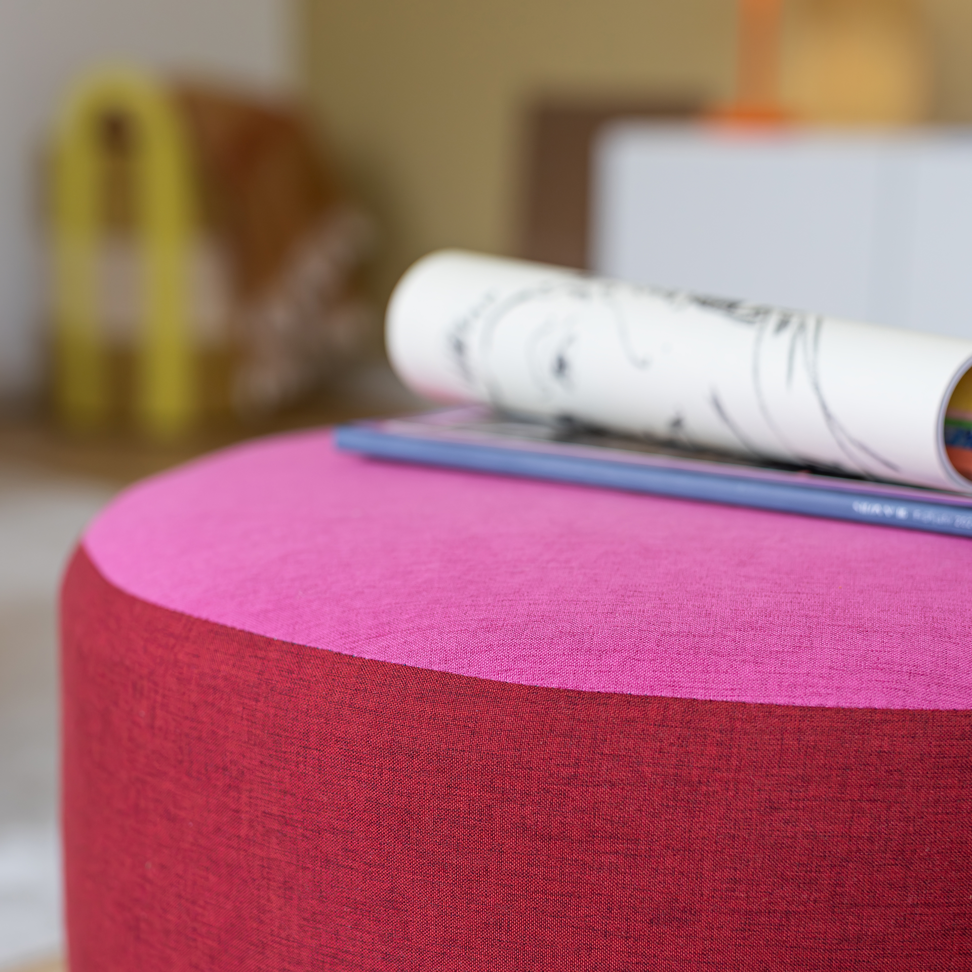 Gros plan sur un pouf rond « Pepe berry » recouvert d’un tissu bicolore, couleur baie foncée sur les côtés et rose vif sur le dessus. Sur l’assise reposent un dessin enroulé et un mince livre bleu. Le pouf sert de support pratique.