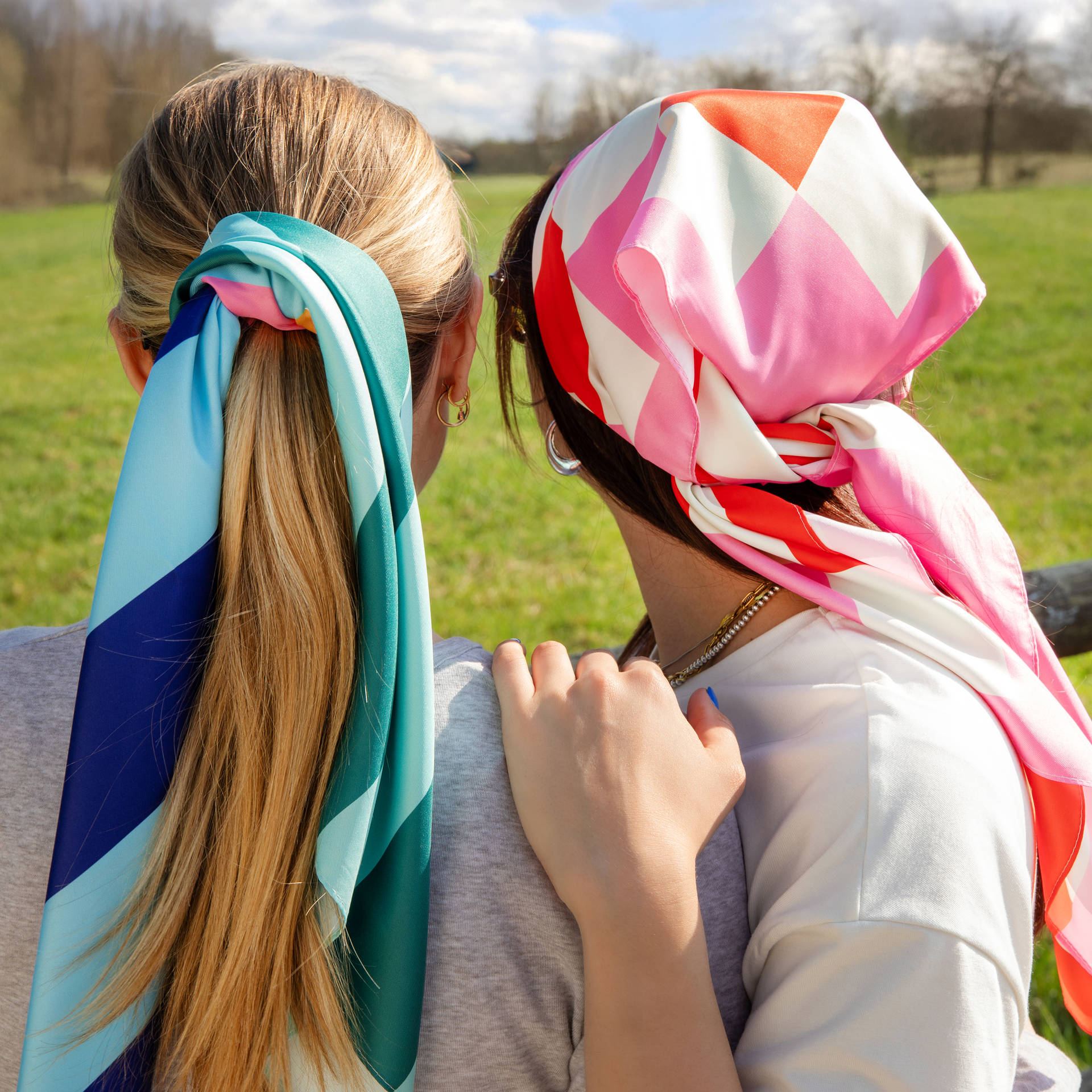 Zwei Frauen von hinten, eine mit Halstuch als Haargummi, die andere mit Halstuch als Kopftuch. Im Hintergrund eine Wiese und Bäume.