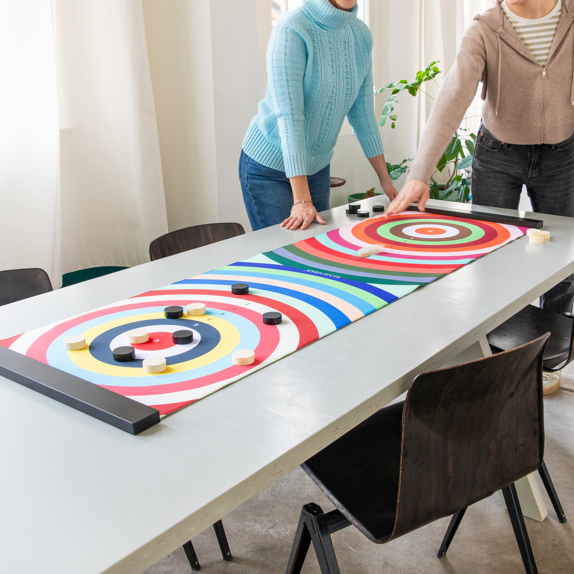 Zwei Personen spielen Colour Curling auf einem Tisch. Das Spielfeld ist bunt mit konzentrischen Kreisen. Schwarze und helle Spielsteine liegen auf dem Feld.