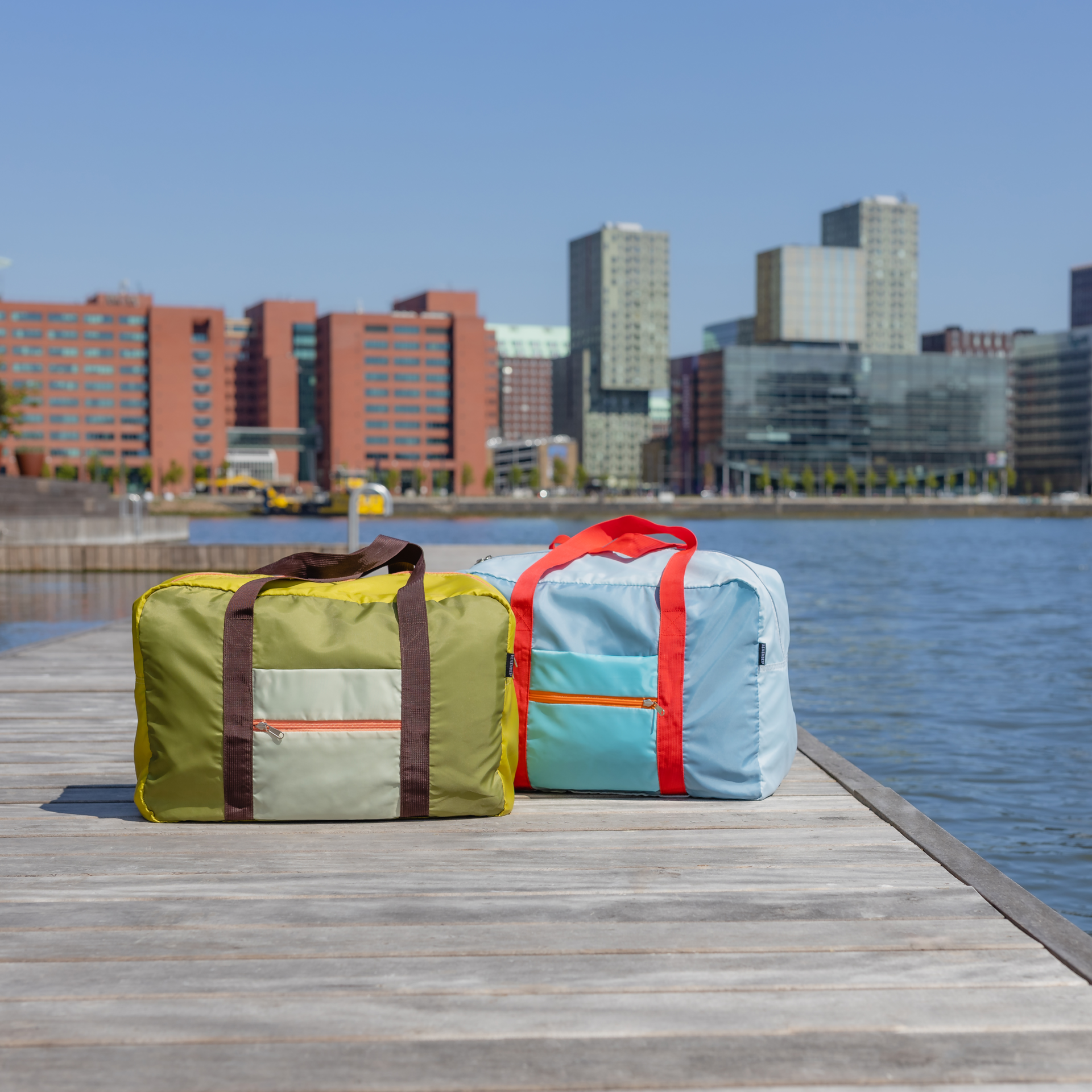 Zwei 'Liz' Reisetaschen, eine hellgrüne und eine hellblaue, stehen auf einem Holzsteg vor einer Skyline mit Wolkenkratzern und blauem Wasser. Die Taschen haben farblich passende Tragegriffe.