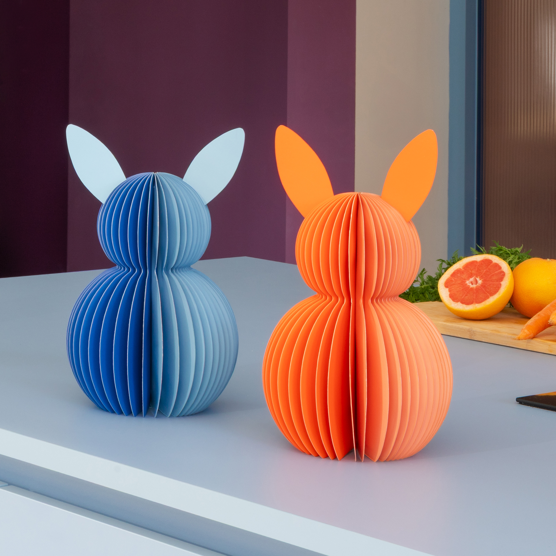 Zwei dekorative Papierhasen, blau und orange, stehen auf einer hellgrauen Oberfläche vor unscharfem Hintergrund mit Obst. Die Hasen sind aus gefächertem Papier.