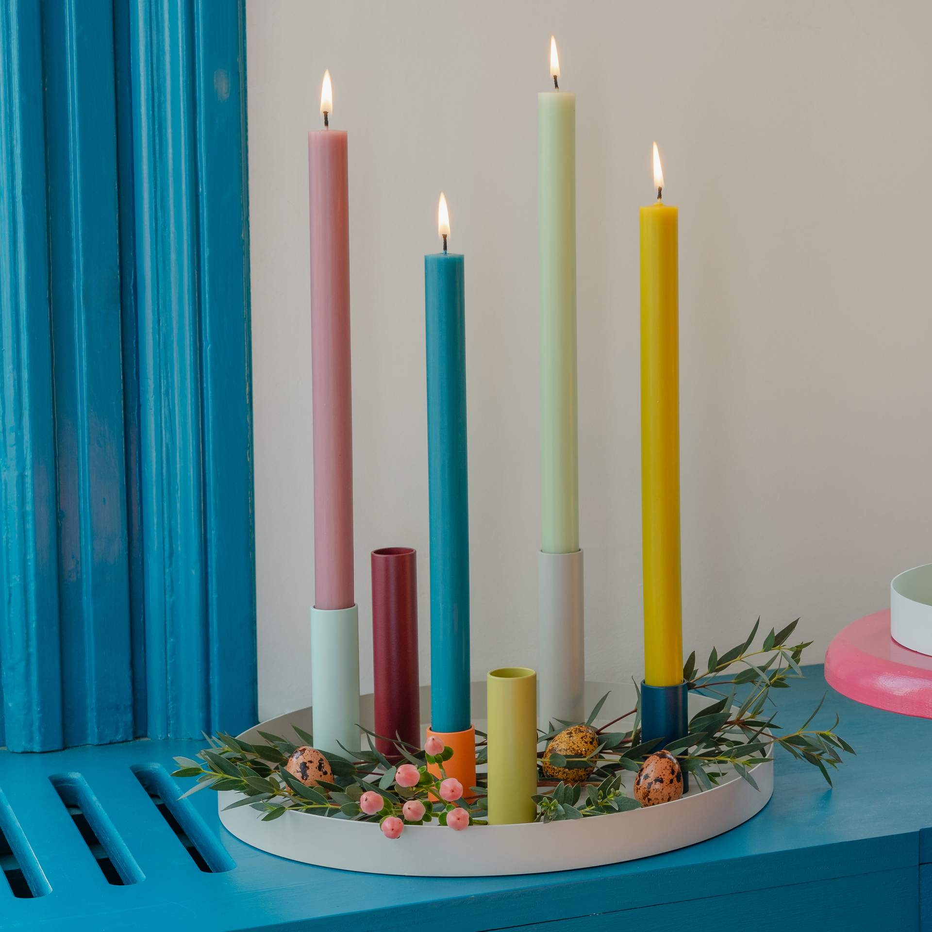 Ein Arrangement aus farbigen Kerzen und passenden Kerzenhaltern auf einem weißen runden Tablett. Verziert mit floralen Elementen und dekorativen Eiern. Einige Kerzen sind angezündet.