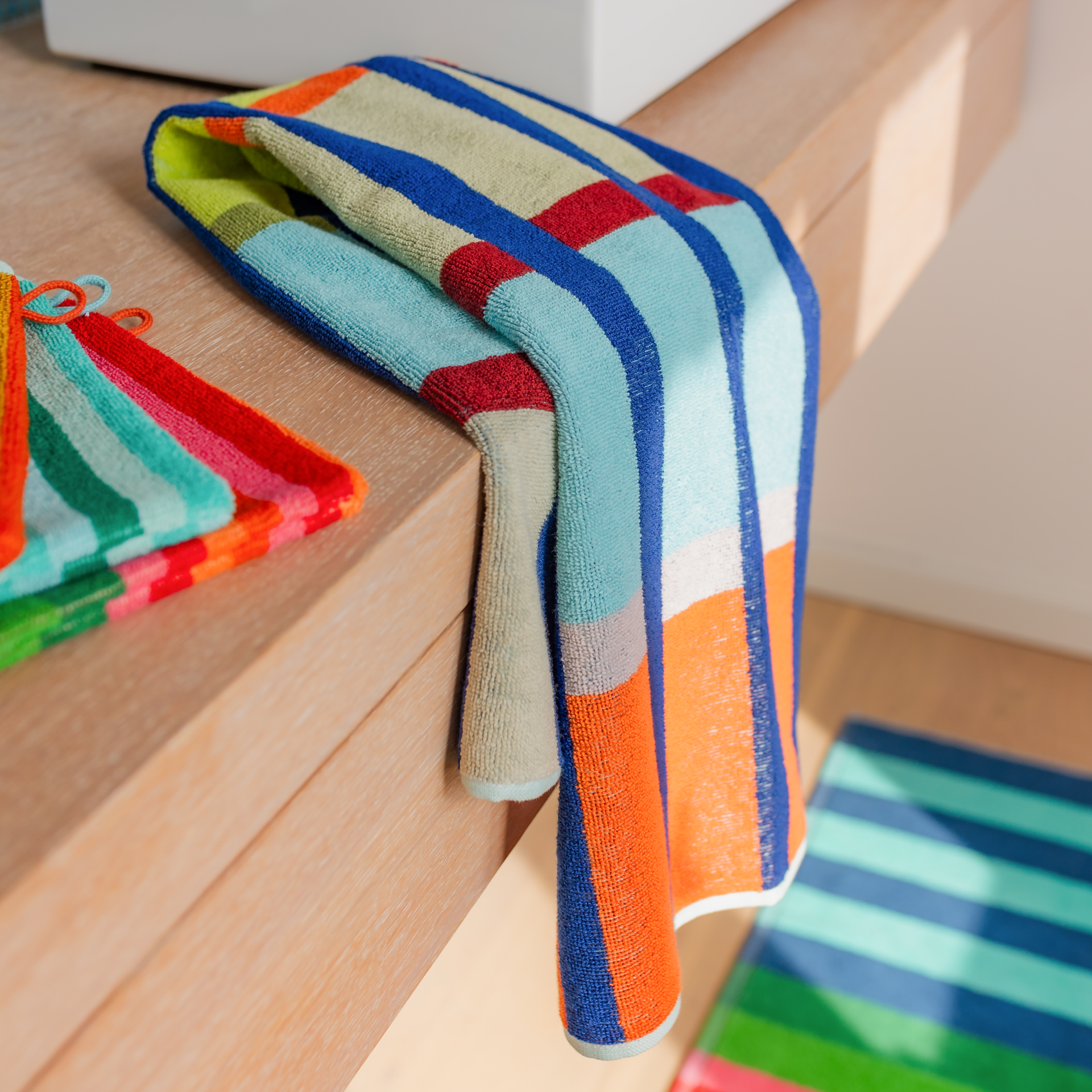 Serviette Yoku pliée verticalement ou à plat, avec motif à carreaux vifs sur fond blanc. Grandes rayures verticales bleu foncé, bandes horizontales bleu clair, jaunes, vertes et corail, avec fines lignes marron et roses.