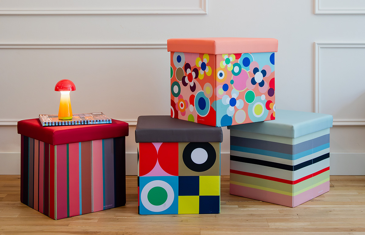 Quatre tabourets colorés avec rangement sur parquet clair, devant un mur blanc. À gauche, un tabouret à rayures rouges avec une lampe champignon rouge et un magazine. Les autres : floral, géométrique, rayures ; dessus uni.