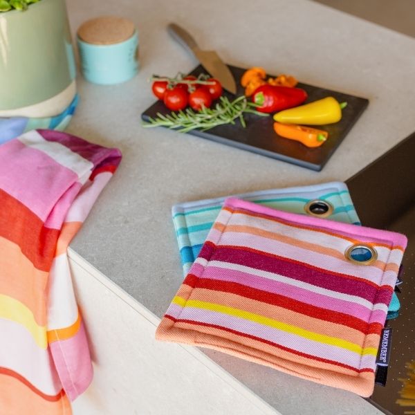 Deux maniques rayées aux couleurs vives et un torchon assorti sont posés sur un plan de travail de cuisine. Sur une planche à découper noire à l'arrière-plan : un couteau, des tomates, du romarin et un poivron. Une plante verte vient compléter le tableau.