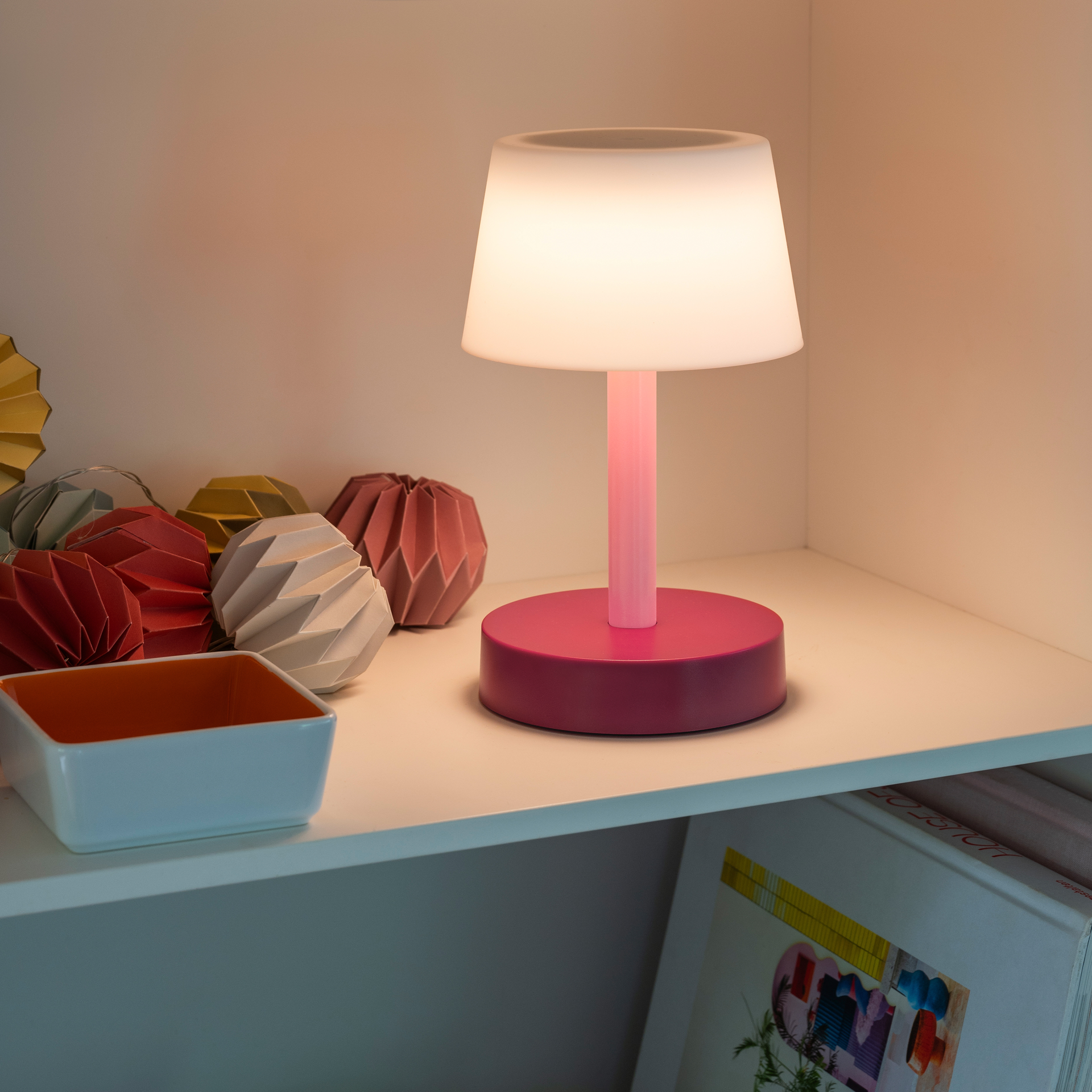 Das Produkt zeigt die Tischleuchte Fritz 'coral' mit aktiviertem, warmweißem Licht auf einem weißen Regal. Dekorative Faltobjekte und eine Schale sind daneben platziert.