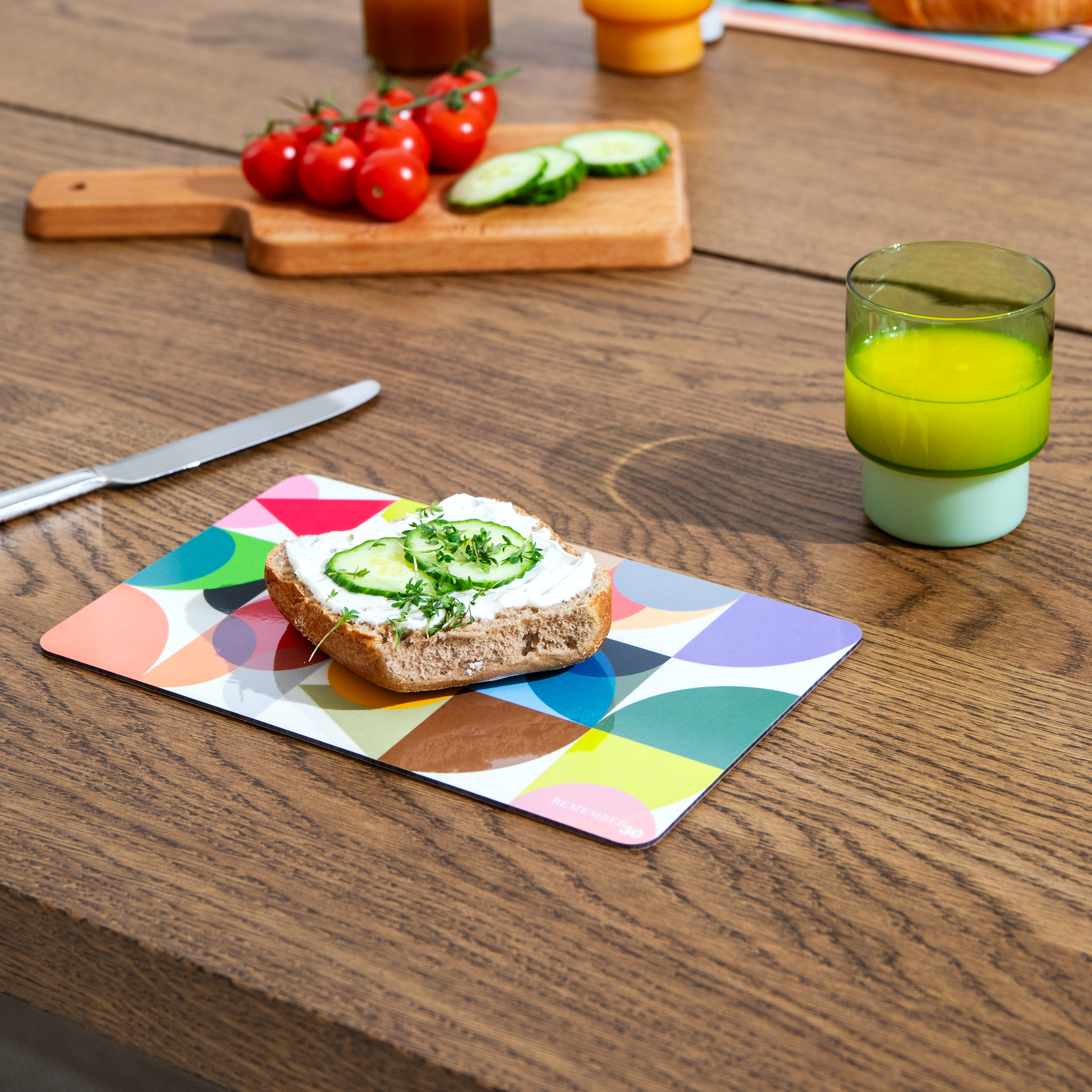 Ein Frühstücksbrettchen 'Remember 30' mit buntem Kreismuster, bestückt mit Brot, Frischkäse, Gurke und Kresse. Daneben ein Messer, Tomaten und ein Glas Saft auf einem Holztisch.