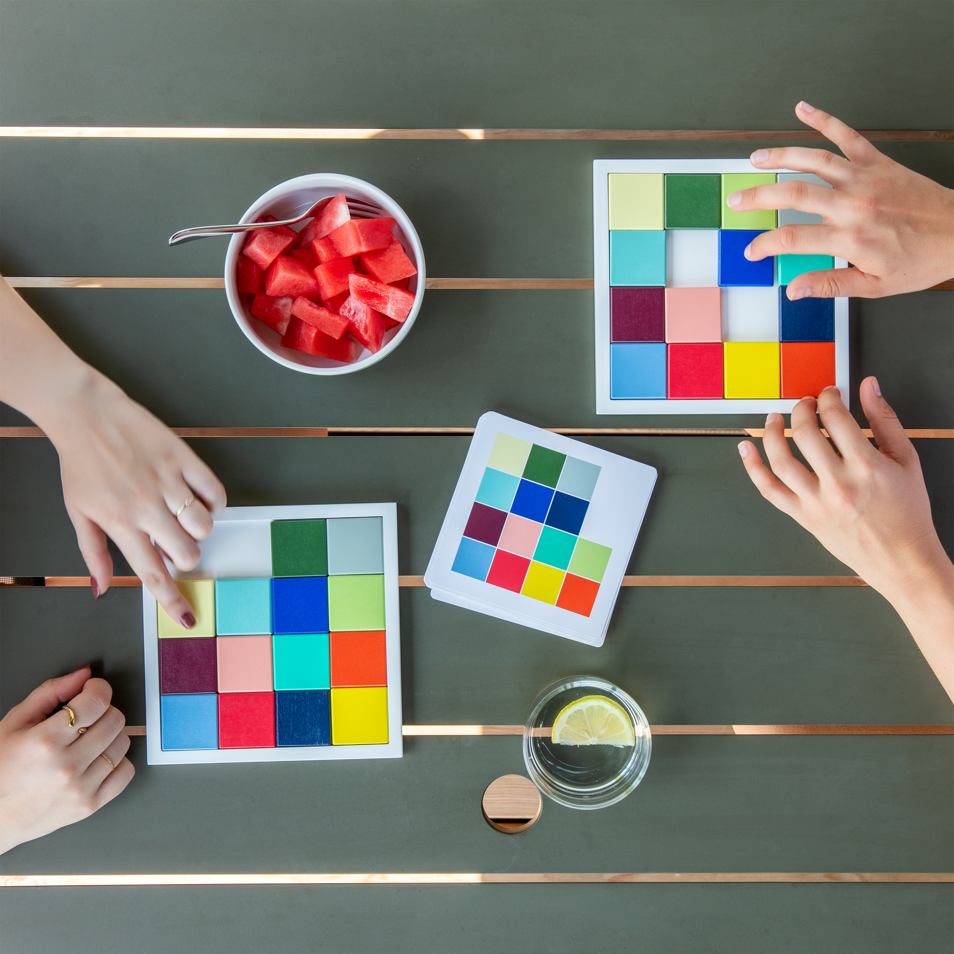 Trois paires de mains sont sur une table en bois vert foncé et jouent avec deux puzzles colorés. Une main pousse une pièce jaune en bas à gauche, une autre une bleue en haut à droite. À côté, un nuancier, une pastèque et de l'eau.
