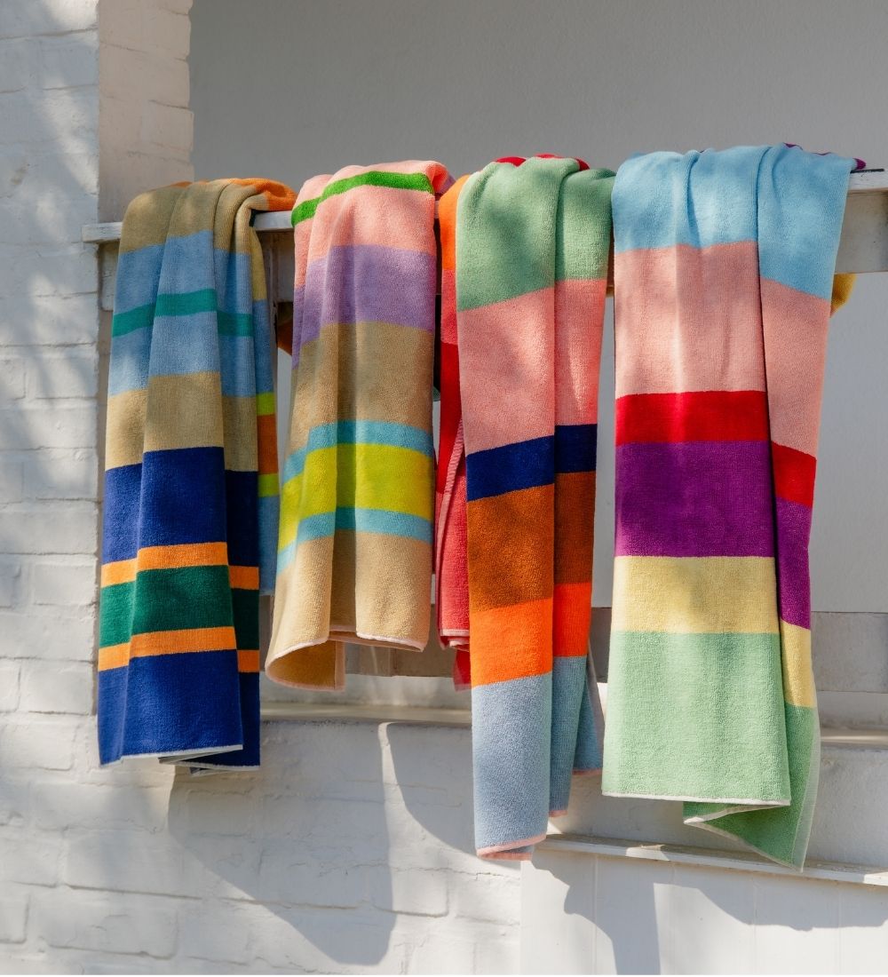 Vier leuchtend bunte Handtücher mit horizontalen Streifen hängen über einer weißen Leiste und trocknen in der Sonne. Sie zeigen kräftige Farben wie Blau, Orange, Grün, Gelb, Rosa, Rot und Violett. Im Hintergrund eine weiße Ziegelwand.