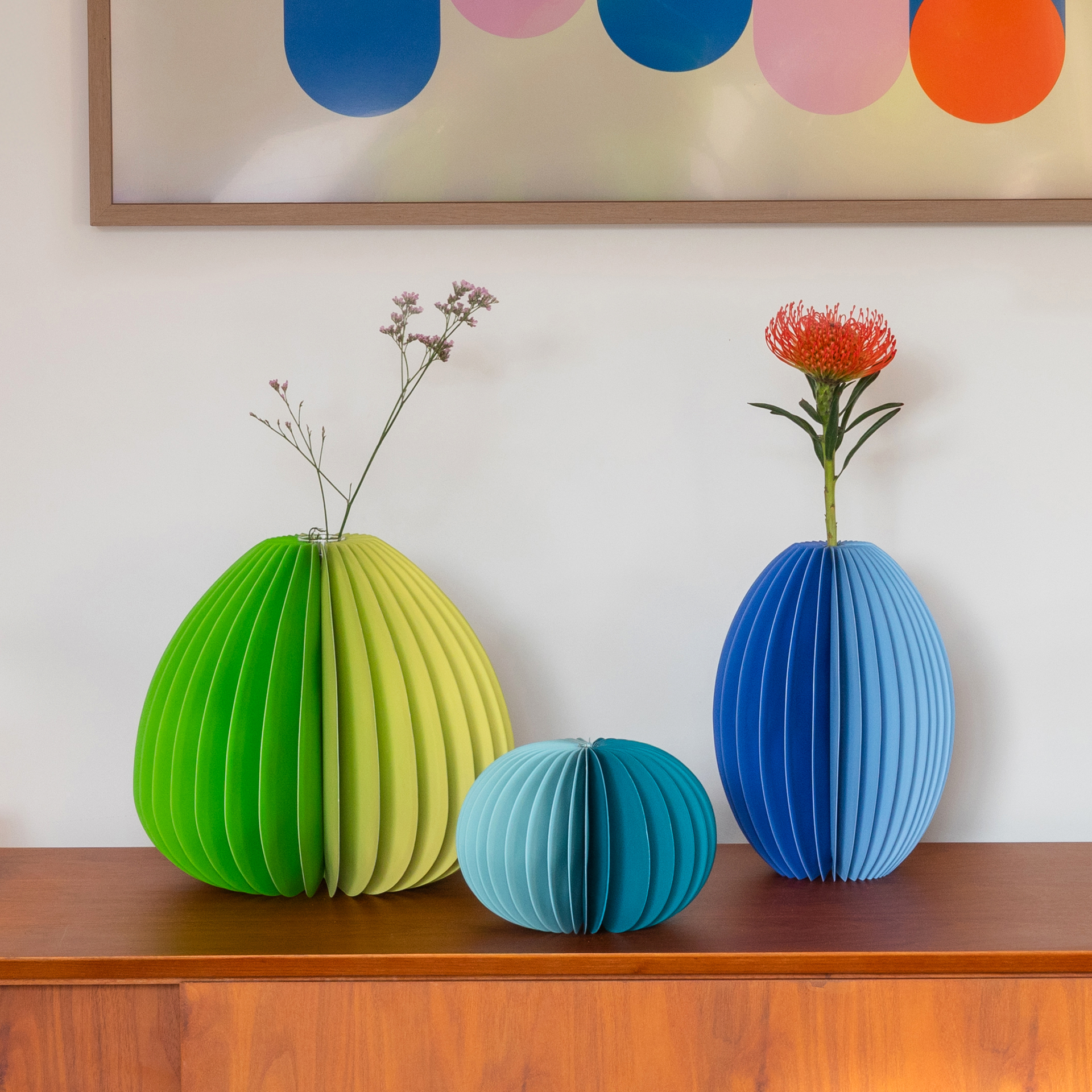 Auf einem Holzschrank stehen drei farbige Papier-Vasen. In der linken grünen Vase ist eine kleine rosa Blüte, in der rechten blauen eine rote. Über ihnen hängt ein Bild mit farbigen Kreisen.