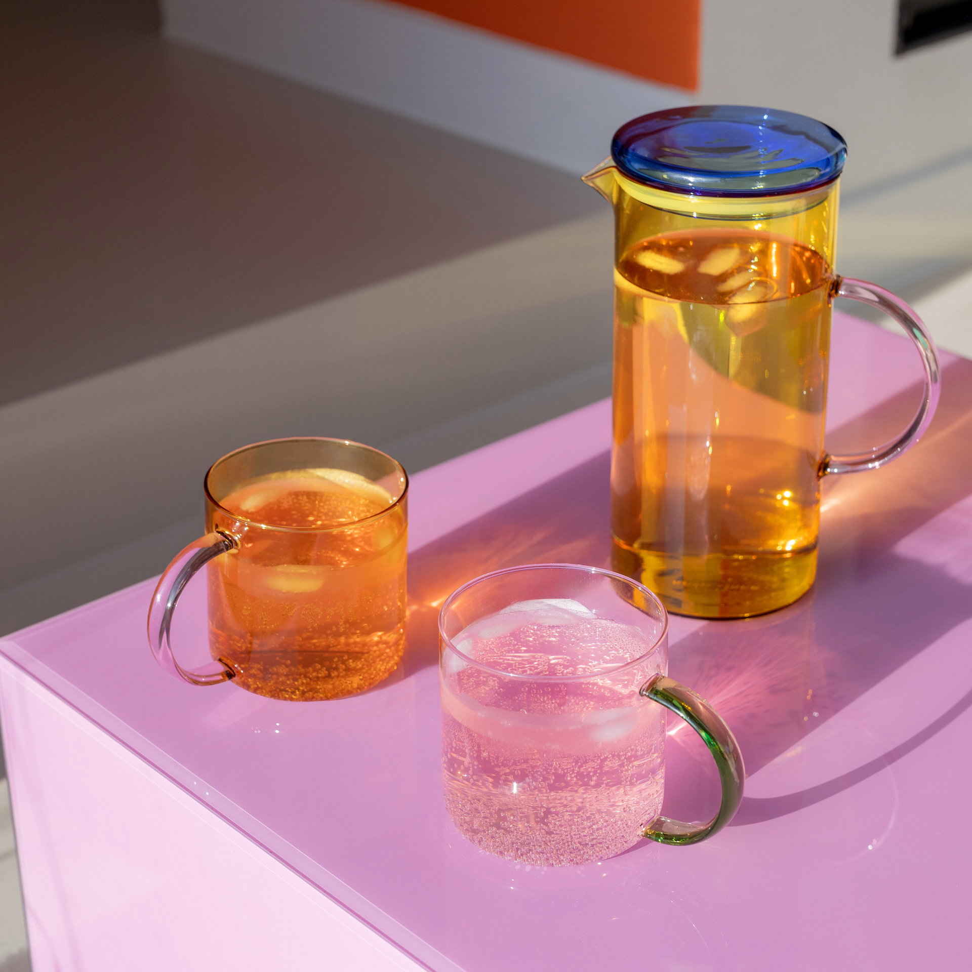 Brocca di vetro gialla con coperchio blu e due bicchieri con bevande su un tavolo rosa.