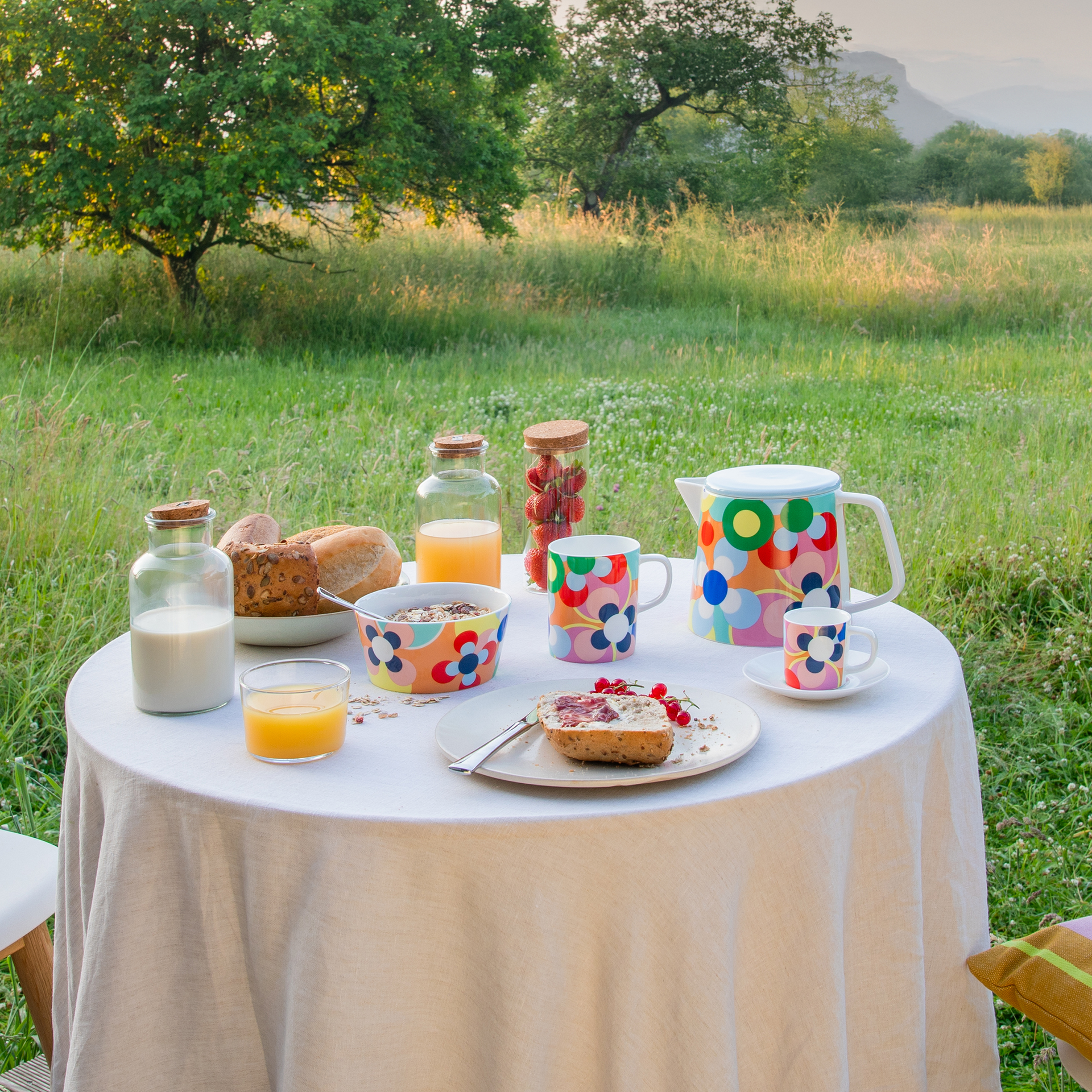 Frühstückstisch im Grünen: Weißes Tischtuch, bunte Tassen/Kanne, Brot, Müsli, Saft, Milch und Erdbeeren. Im Hintergrund Wiese, Baum und Berge.