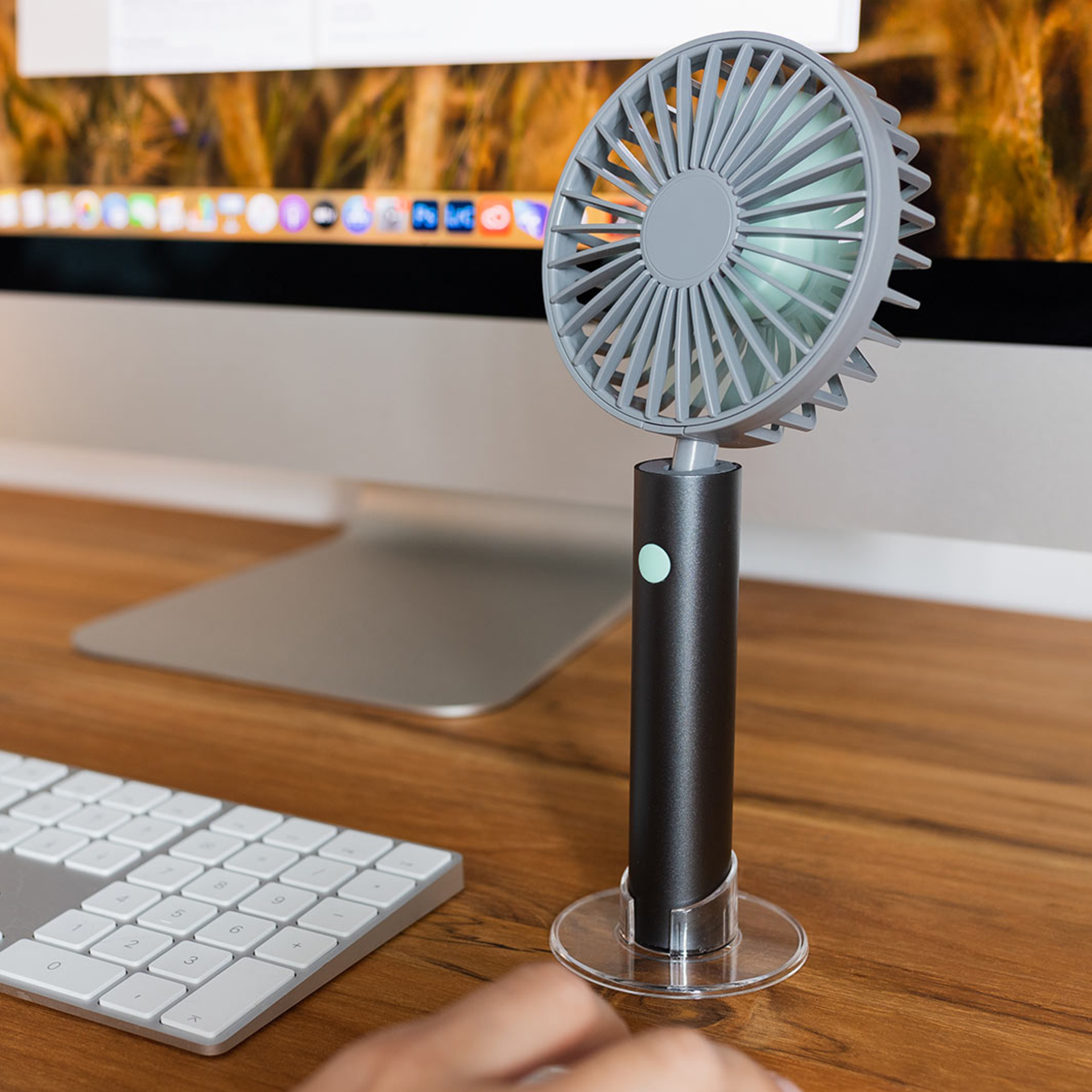 Un ventilatore Flow color grafite con base trasparente si trova su una scrivania accanto a una tastiera e a un monitor.