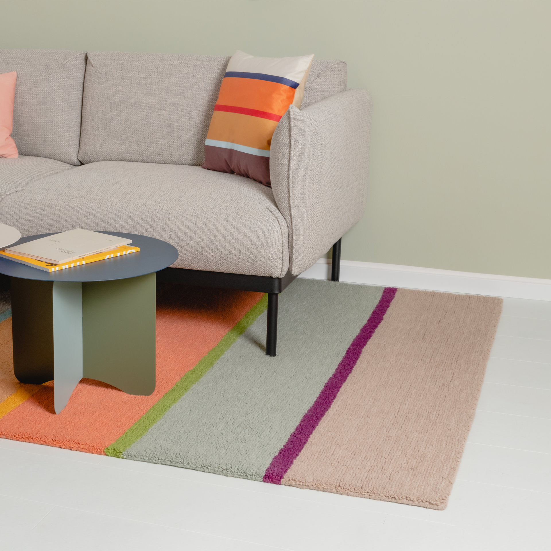 Tappeto 'Cambridge' in Ambiente: vista parziale del divano, del tavolino e del tappeto a strisce colorate in arancione, verde, viola e beige su un pavimento in legno chiaro.