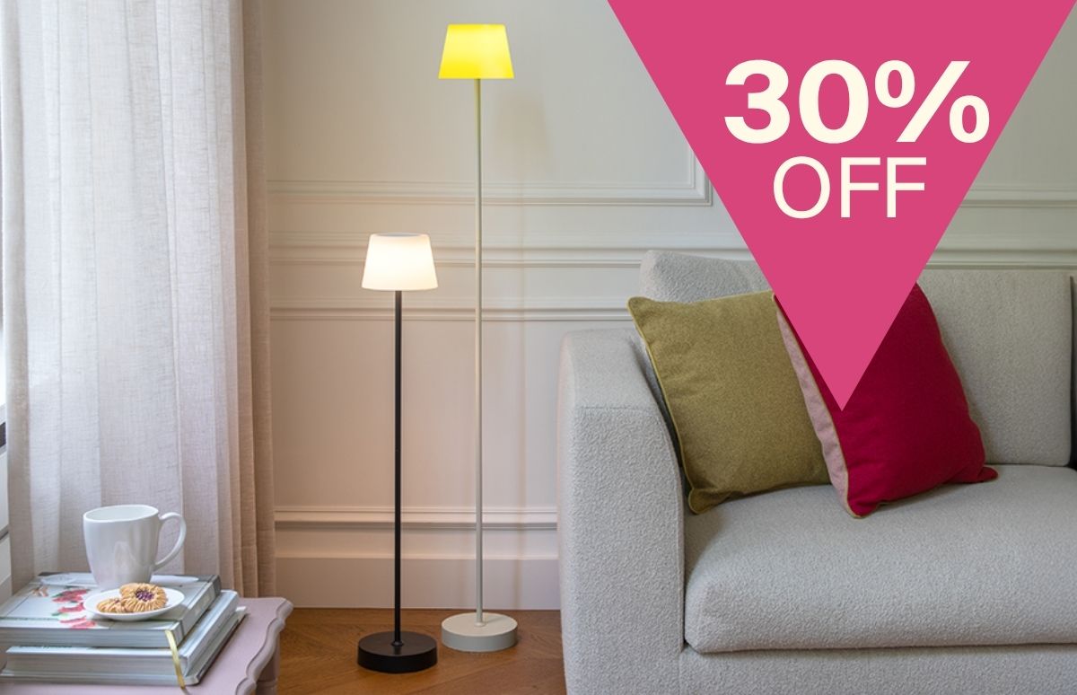 Zwei moderne Stehlampen in elegantem Wohnzimmer. Schwarze Lampe mit weißem, weiße mit gelbem Schirm leuchten. Links Bücher, Tasse, Kekse auf Tisch. Rechts beiges Sofa mit olivgrünem und rotem Kissen. Pinkes „30% OFF“-Dreieck oben rechts.