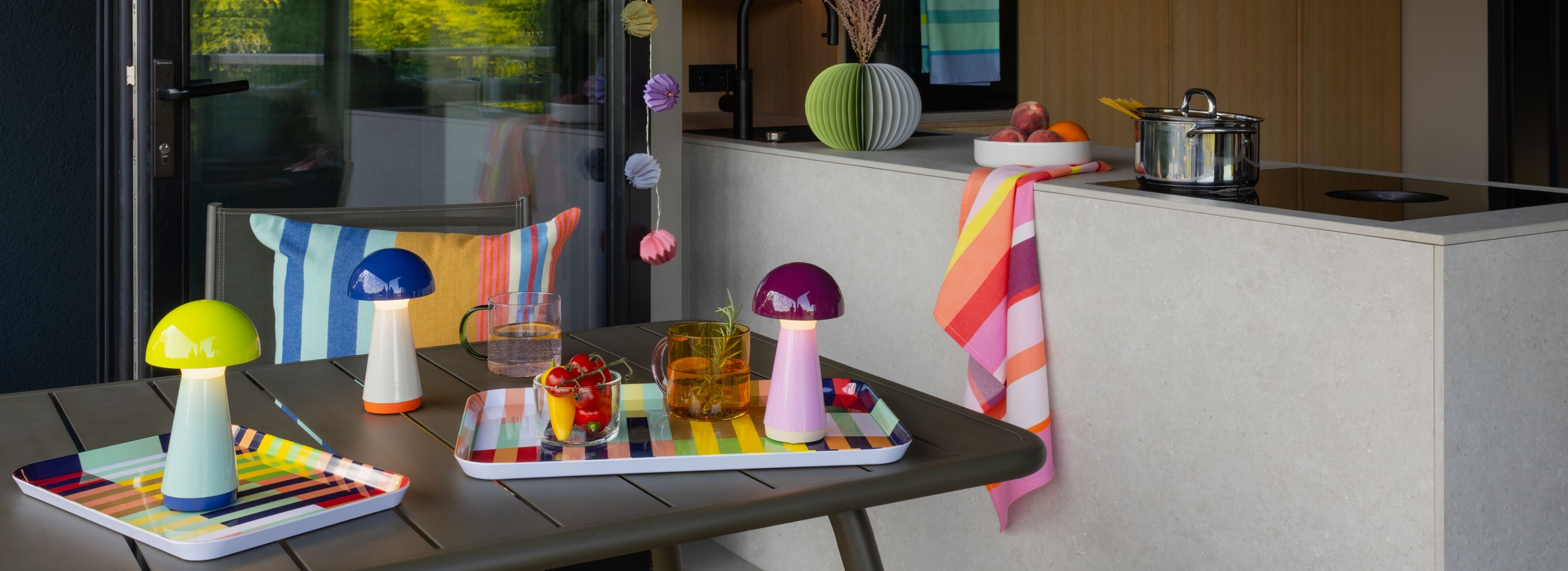 Trois lampes champignon aux couleurs vives sur une table d'extérieur sombre, près de deux plateaux rayés avec boissons et légumes frais. En arrière-plan, cuisine moderne, casserole de pâtes et corbeille de fruits.