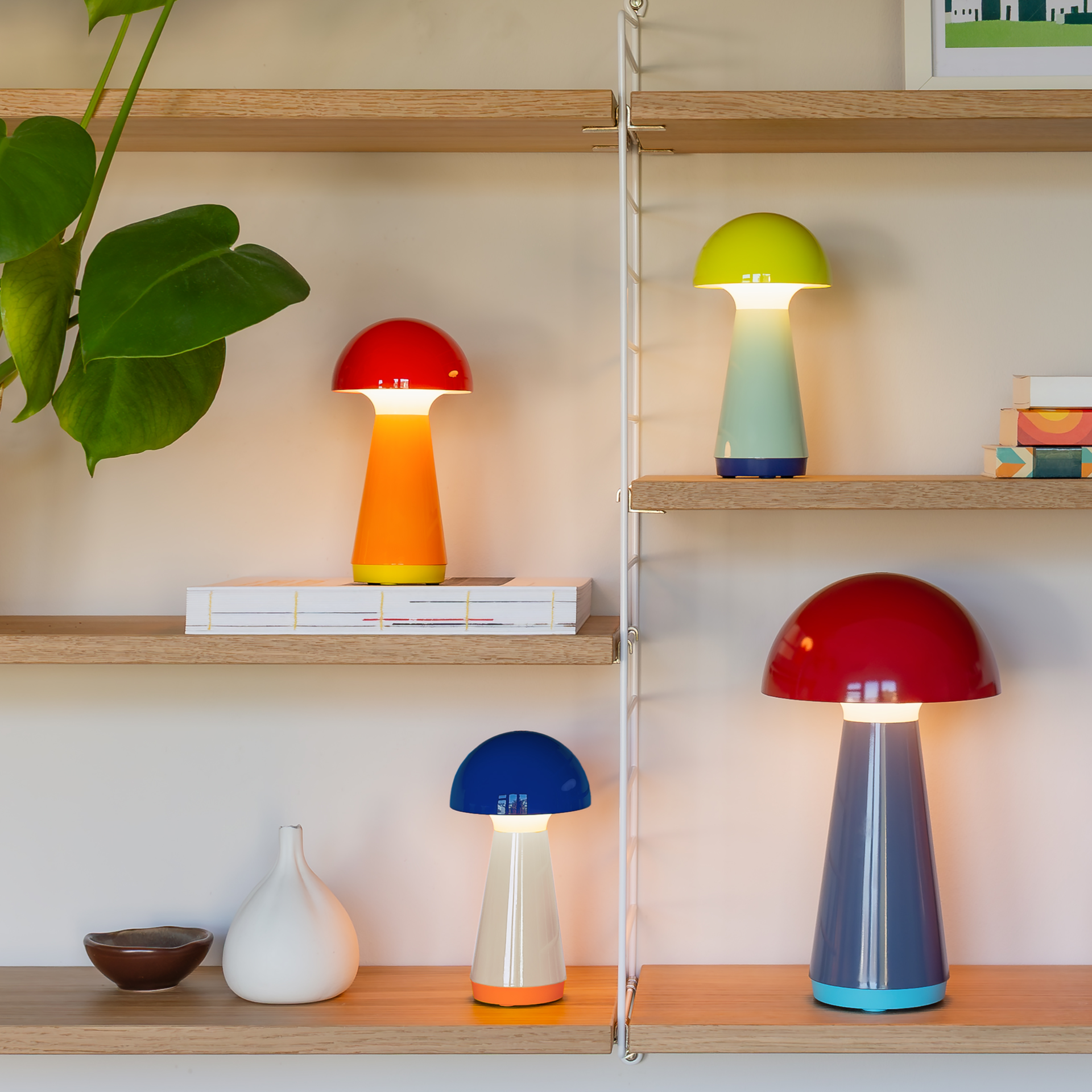 Regal mit vier pilzförmigen Lampen in Rot, Grün und Blau, zwei Vasen, Büchern und einer grünen Pflanze.
