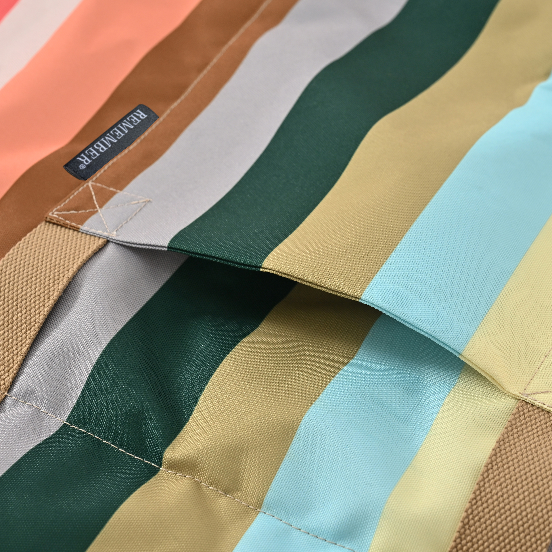 Gros plan sur une couverture colorée pour chien d’extérieur, à rayures vives pêche, marron, gris clair, vert foncé, olive, bleu clair et jaune. Tissu robuste à texture tissée, coutures et étiquette « REMEMBER® » cousue.