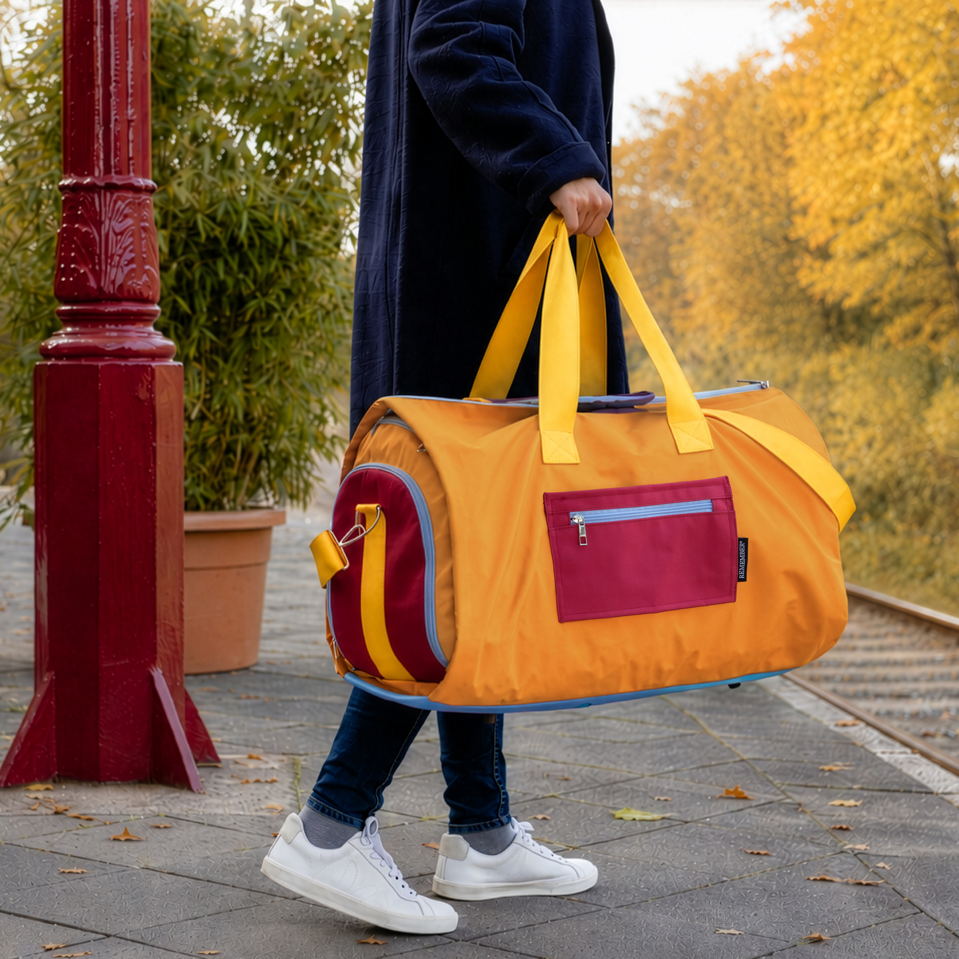 Sac de voyage Lennard Pumpkin de couleur orange, doté de poignées jaunes et d'une bandoulière rembourrée bordeaux. Des détails de fermeture éclair bleu clair et des extrémités bordeaux soulignent le design. En matière hydrofuge.