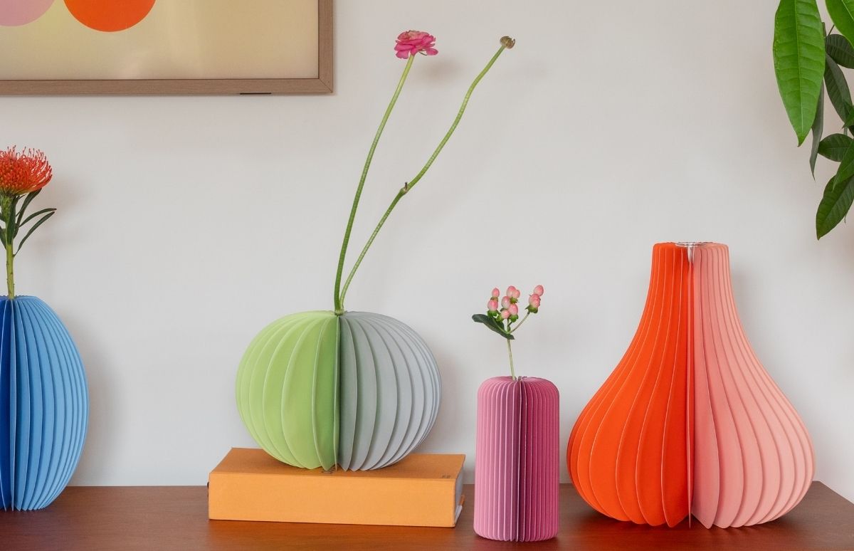 Vier gefaltete Papier-Vasen in lebhaften Farben auf Holz: blaue Kugelvase mit roter Blüte, grüne und graue Kugelvase auf orange Box mit rosa Ranunkel. Daneben orange-rosa Birnenvase und pinke Zylindervase mit Beeren. Weißer Hintergrund.